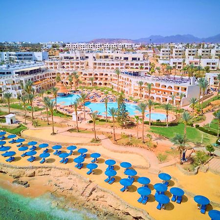 Pickalbatros Royal Grand Sharm - Adults Friendly 16 Years Plus Exterior photo The photo shows a vibrant resort area with several buildings that appear to be accommodations or hotel units. There are multiple swimming pools surrounded by palm trees, creating a tropical atmosphere. In the foreground, there is a beach area with sa