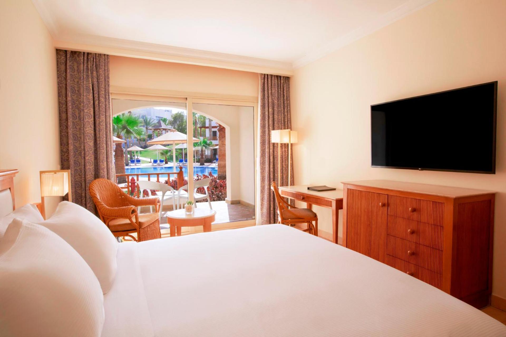 Pickalbatros Royal Grand Sharm - Adults Friendly 16 Years Plus Exterior photo The photo shows a hotel room featuring a large bed with white bedding. There are wooden furniture pieces, including a dresser and a desk. A flat-screen television is mounted on the wall opposite the bed. The room has large windows with sheer curtains