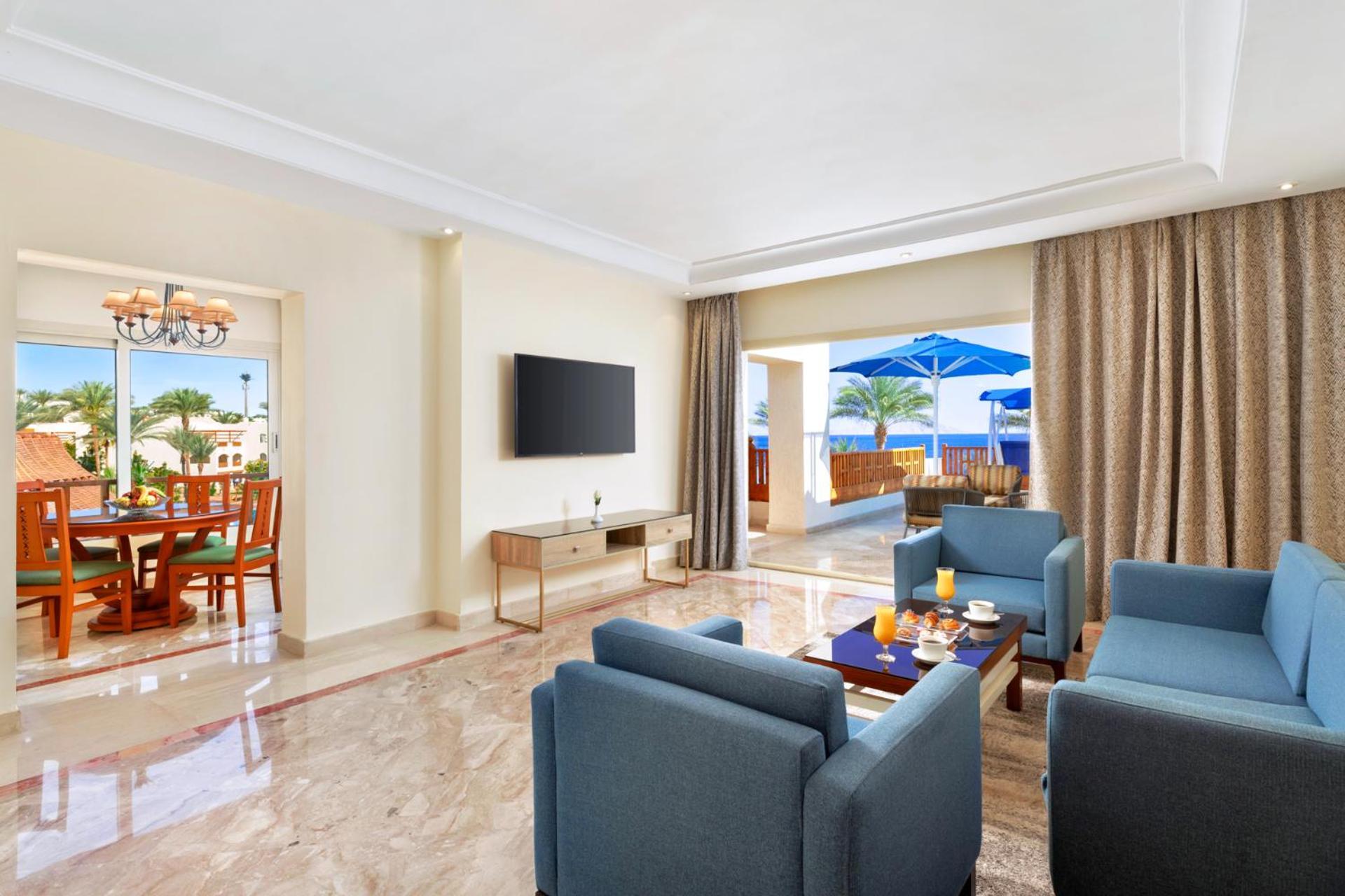 Pickalbatros Royal Grand Sharm - Adults Friendly 16 Years Plus Exterior photo The photo shows a modern and spacious living area. There are several blue chairs arranged around a coffee table, which holds some refreshments. A large flat-screen TV is mounted on the wall, and there are sliding glass doors that open to an outdoor a