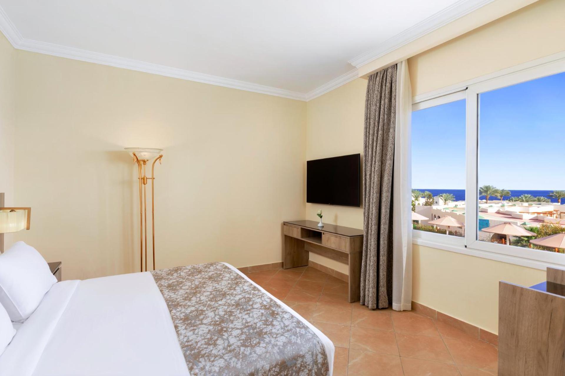 Pickalbatros Royal Grand Sharm - Adults Friendly 16 Years Plus Exterior photo The photo shows a hotel room with a modern and minimalistic design. There is a comfortable bed with white linens and a decorative gray throw at the foot. A large window offers a view of the outdoors, possibly with a glimpse of the sea or beach. A fla