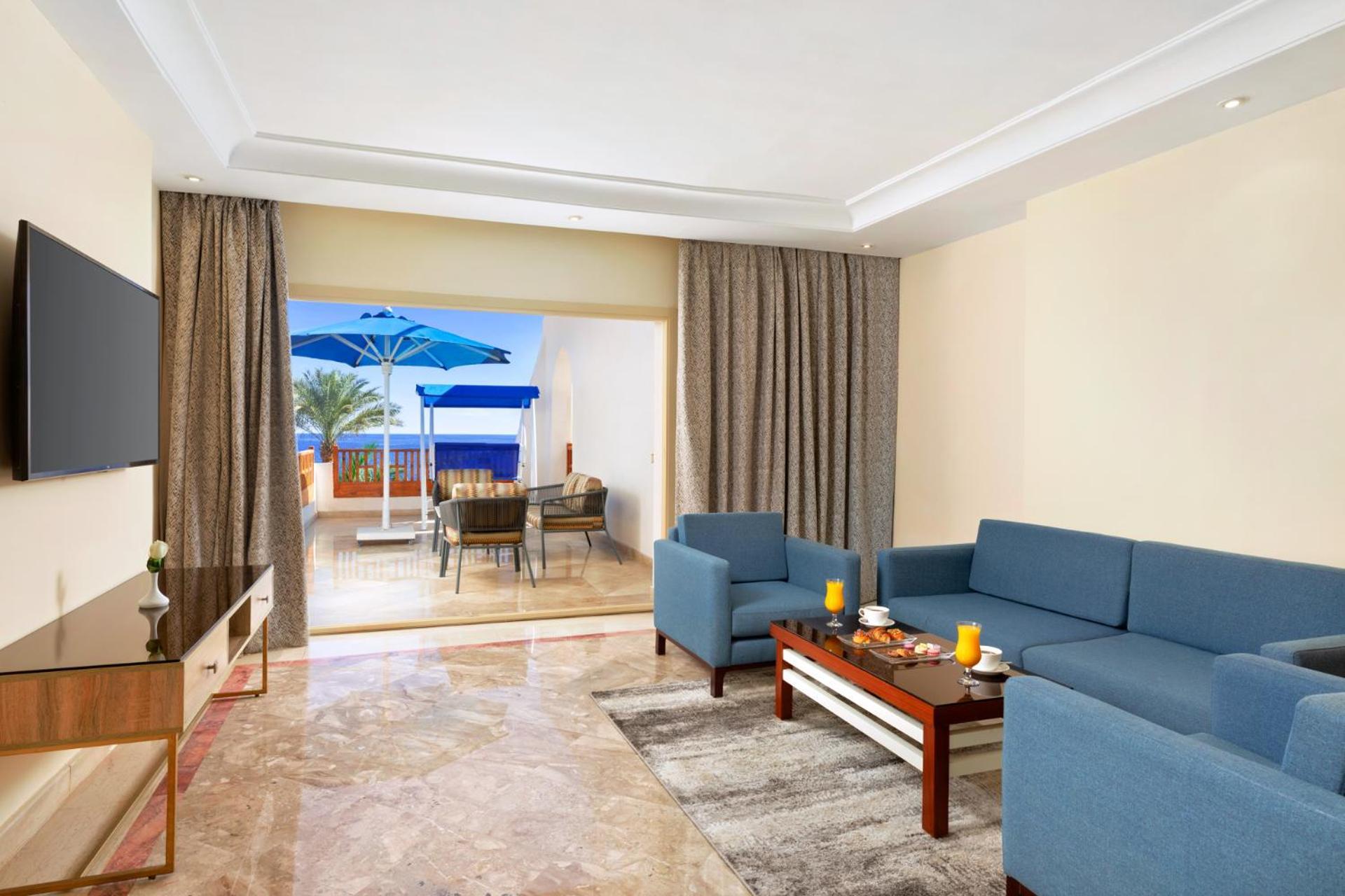 Pickalbatros Royal Grand Sharm - Adults Friendly 16 Years Plus Exterior photo The photo shows a spacious living area with modern decor. There are two blue sofas arranged around a coffee table that holds some decorative items and candles. A flat-screen TV is mounted on the wall across from the seating area. To the left, there a