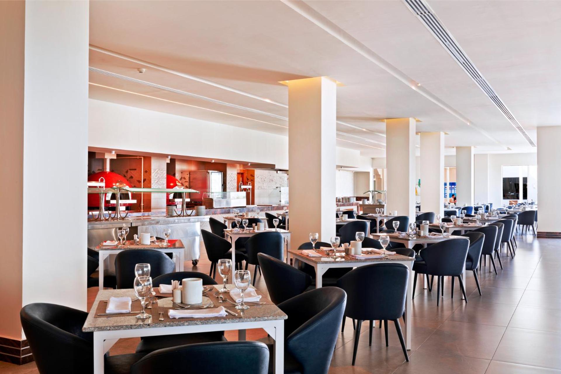 Pickalbatros Royal Grand Sharm - Adults Friendly 16 Years Plus Exterior photo The photo shows a modern restaurant interior. It features a spacious layout with several dining tables arranged in a row, each set with dishes, glasses, and cutlery. The tables have light-colored surfaces and are accompanied by black chairs. In the b