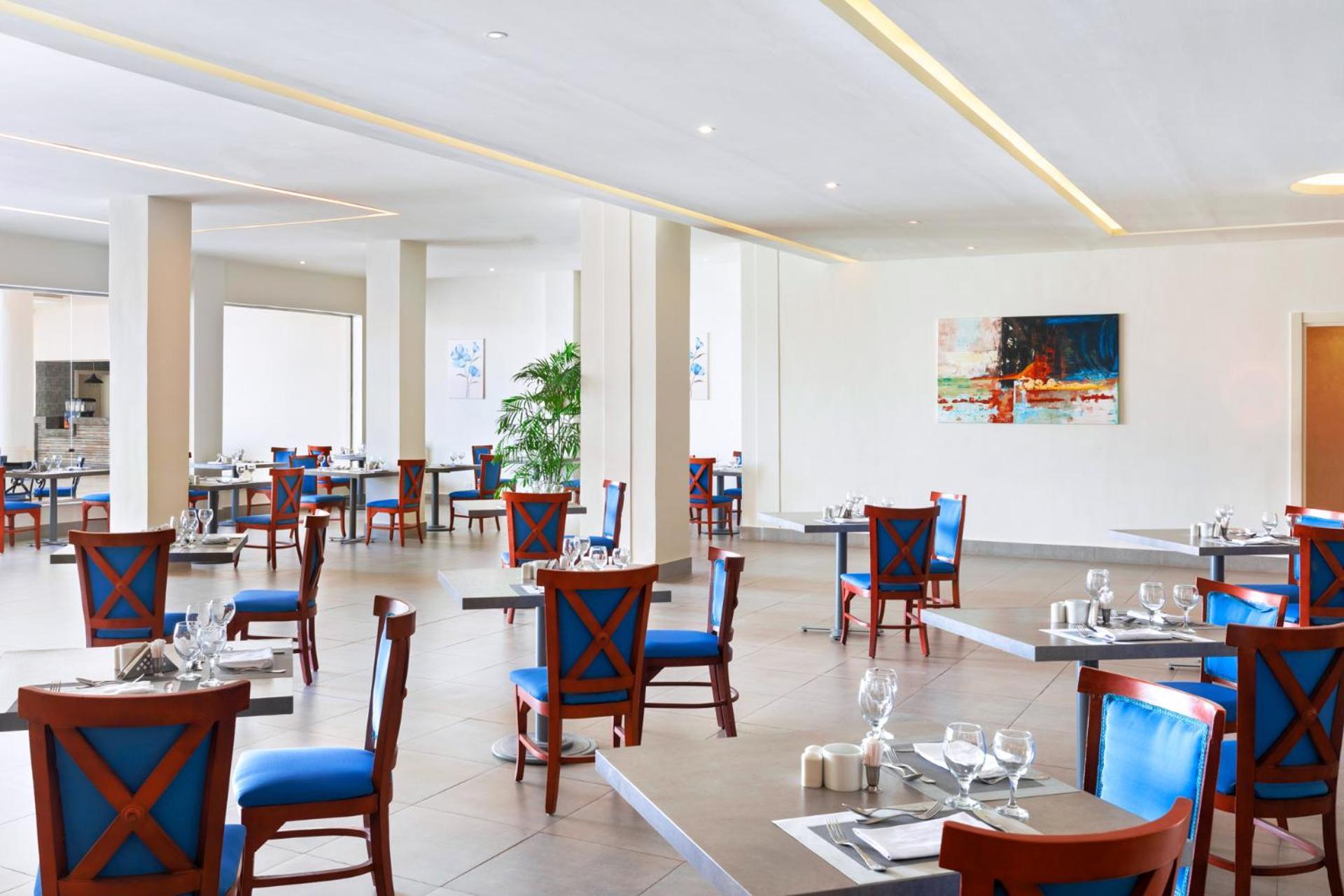 Pickalbatros Royal Grand Sharm - Adults Friendly 16 Years Plus Exterior photo The photo shows an interior of a restaurant featuring a modern and spacious design. The floors are tiled, and the walls are painted in neutral tones. The seating consists of wooden chairs with blue cushions arranged around tables, which are set with 