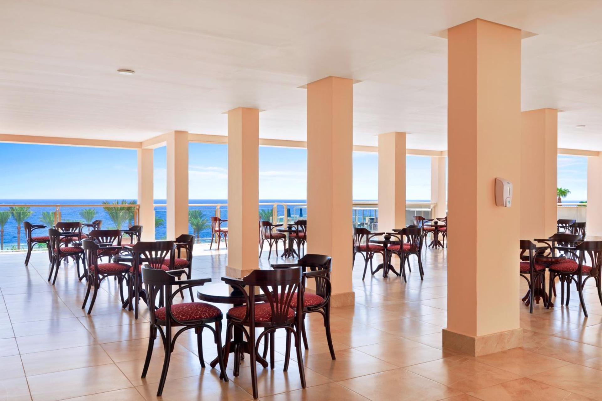 Pickalbatros Royal Grand Sharm - Adults Friendly 16 Years Plus Exterior photo The image shows a dining area or café with several round wooden tables and chairs, featuring red upholstery. The space is well-lit, with large windows that allow a view of the ocean in the background, contributing to a bright and open atmosphere. The