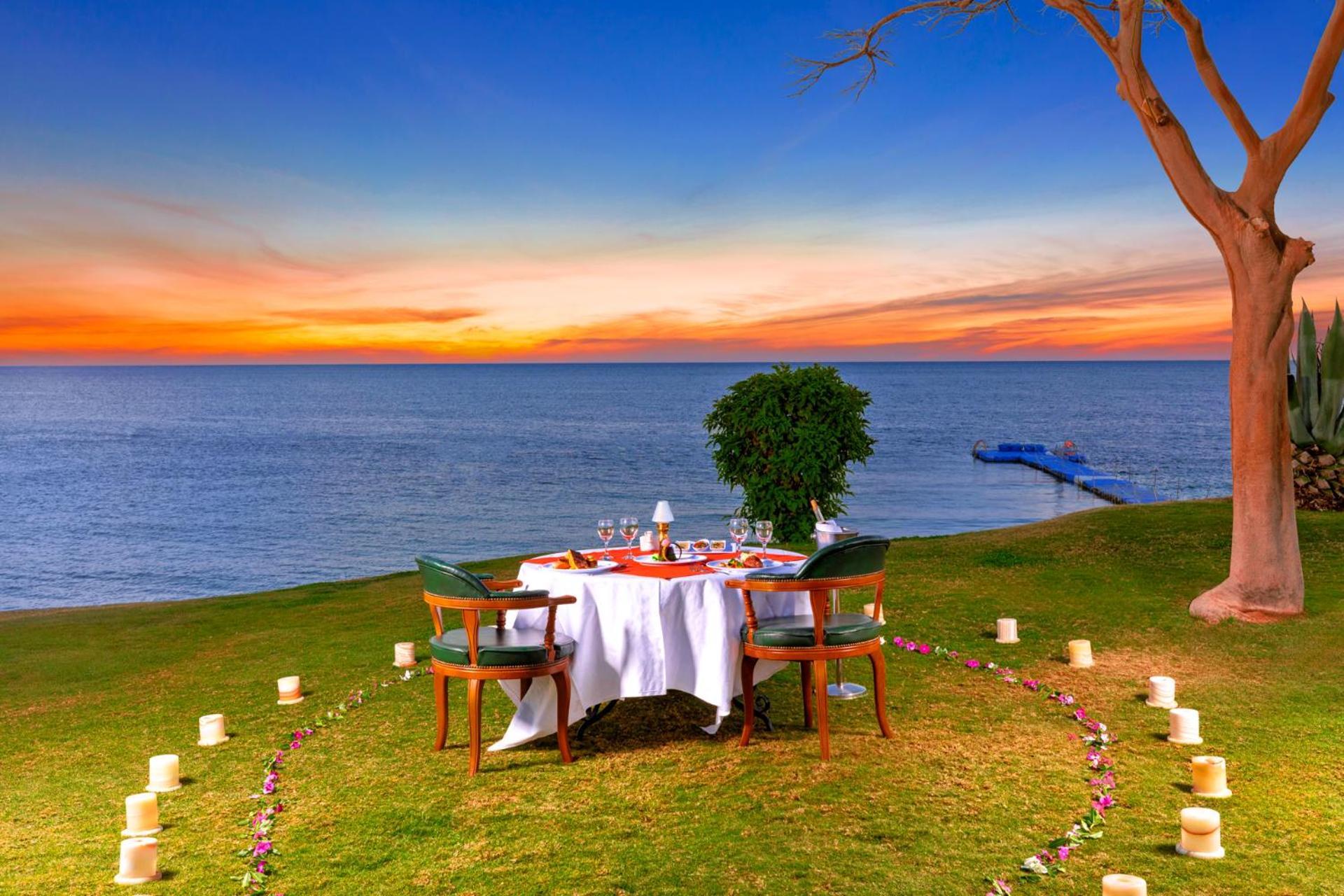 Pickalbatros Royal Grand Sharm - Adults Friendly 16 Years Plus Exterior photo The photo depicts a romantic outdoor dining setup by the seaside. In the foreground, there is a round table elegantly set with a white tablecloth, plates, and glassware, creating a cozy atmosphere. Surrounding the table are green chairs. A circle of 