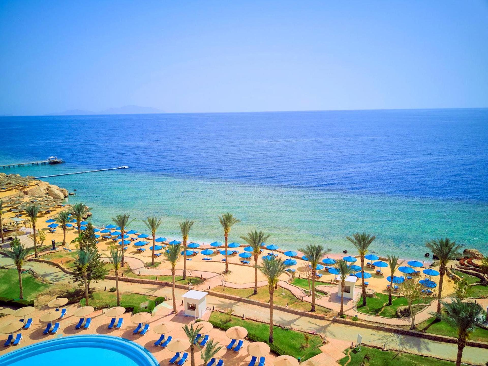 Pickalbatros Royal Grand Sharm - Adults Friendly 16 Years Plus Exterior photo The photo displays a beautiful coastal scene featuring a clear blue sea that transitions into green tones near the shore. In the foreground, there are sandy beaches lined with palm trees and a variety of beach umbrellas. There is also a swimming pool