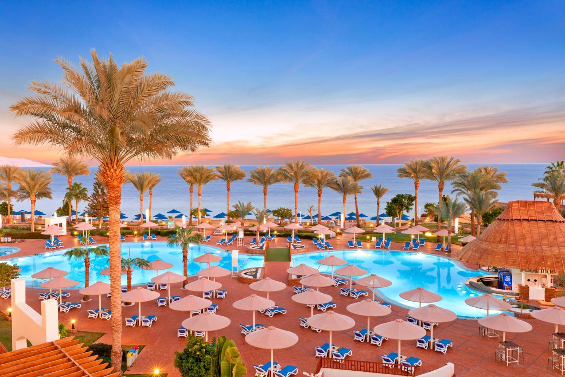 Pickalbatros Royal Grand Sharm - Adults Friendly 16 Years Plus Exterior photo The image shows a beautiful resort setting by the beach. There is a large swimming pool at the center, surrounded by lounge chairs and umbrellas. Palm trees line the area, giving it a tropical vibe. In the background, you can see the ocean and a colo