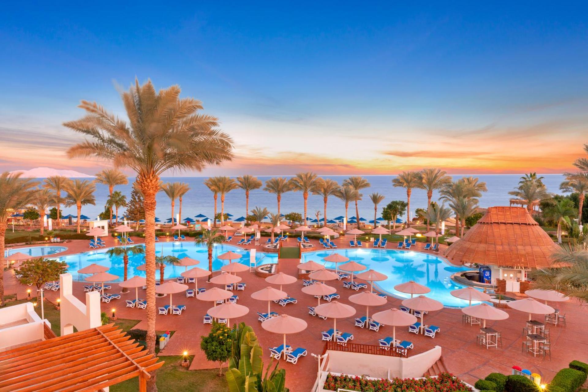 Pickalbatros Royal Grand Sharm - Adults Friendly 16 Years Plus Exterior photo The photo depicts a scenic view of a resort area featuring a large swimming pool surrounded by numerous lounge chairs and umbrellas. In the background, there are tall palm trees and a beautiful sunset over the horizon, creating a vibrant sky. The atm