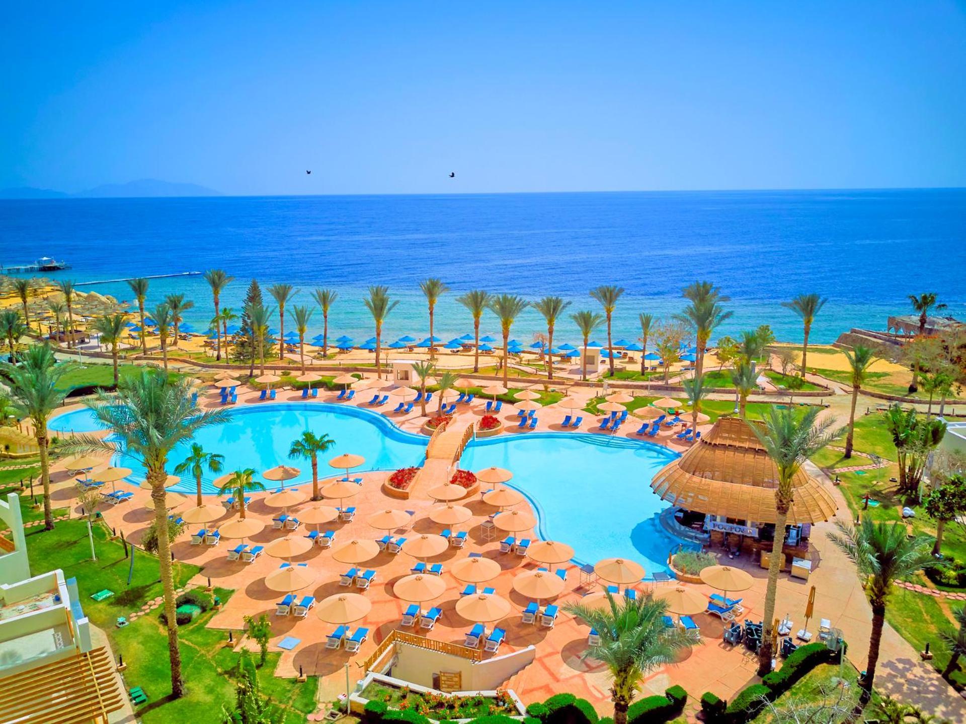 Pickalbatros Royal Grand Sharm - Adults Friendly 16 Years Plus Exterior photo The photo depicts a picturesque resort scene by the sea. In the foreground, there is a large, inviting swimming pool surrounded by lounge chairs and umbrellas. Palm trees are strategically placed around the pool area, adding to the tropical atmospher
