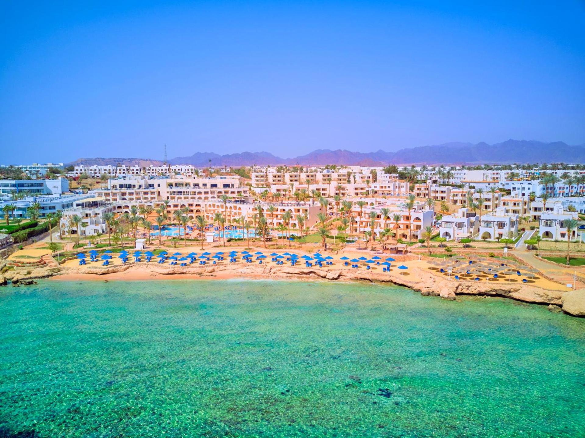 Pickalbatros Royal Grand Sharm - Adults Friendly 16 Years Plus Exterior photo The photo depicts a picturesque beachfront resort. It shows a clear turquoise sea with a sandy shore lined with beach umbrellas and sun loungers. Behind the beach, there are several white buildings of the resort, surrounded by lush greenery. In the b
