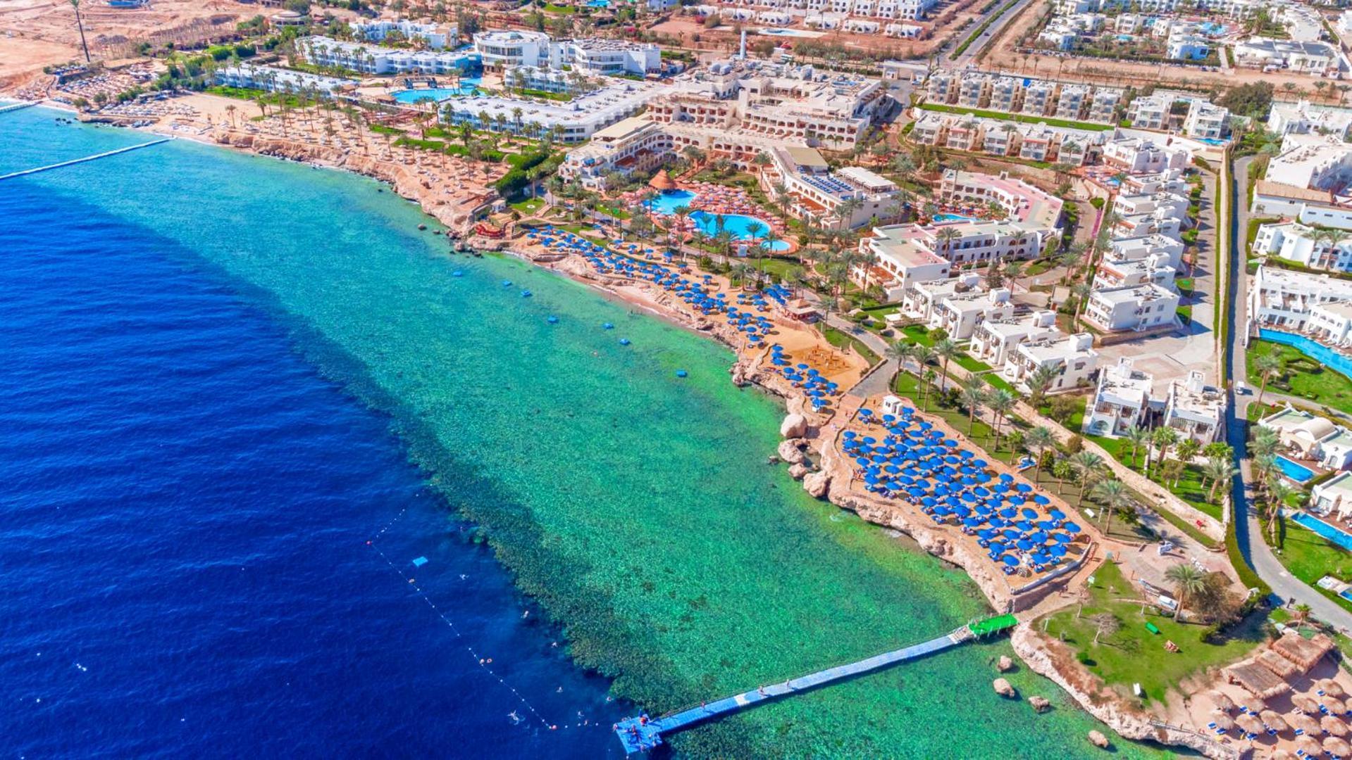Pickalbatros Royal Grand Sharm - Adults Friendly 16 Years Plus Exterior photo The photo showcases a beautiful aerial view of a coastal resort. In the foreground, there's a sandy beach with beach umbrellas and lounge chairs arranged neatly, indicating a tourist area. The clear, turquoise water transitions from the sandy shore t