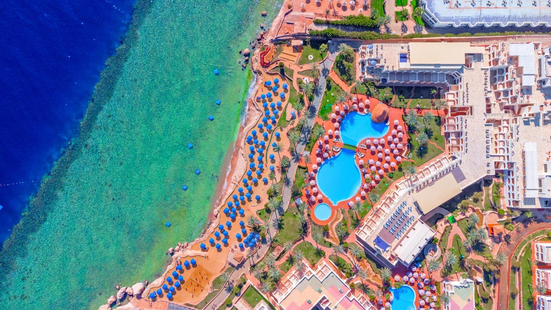 Pickalbatros Royal Grand Sharm - Adults Friendly 16 Years Plus Exterior photo The photo showcases an aerial view of a resort area by the sea. It features a turquoise body of water alongside a sandy beach. There are several umbrellas set up on the beach and lounging areas. A large pool is visible, characterized by its unique sh