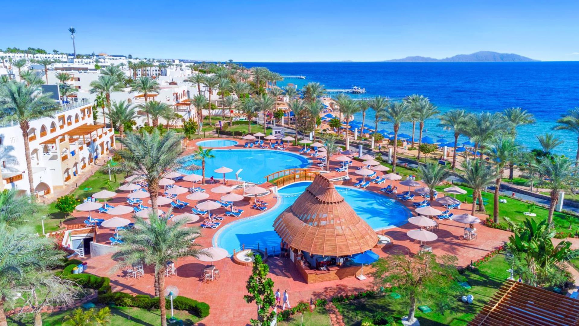 Pickalbatros Royal Grand Sharm - Adults Friendly 16 Years Plus Exterior photo The image depicts a vibrant resort scene overlooking a clear blue sea. In the foreground, there are multiple swimming pools surrounded by sun loungers and umbrellas. A thatched-roof gazebo is situated near the center of the pool area. Palms and lush 