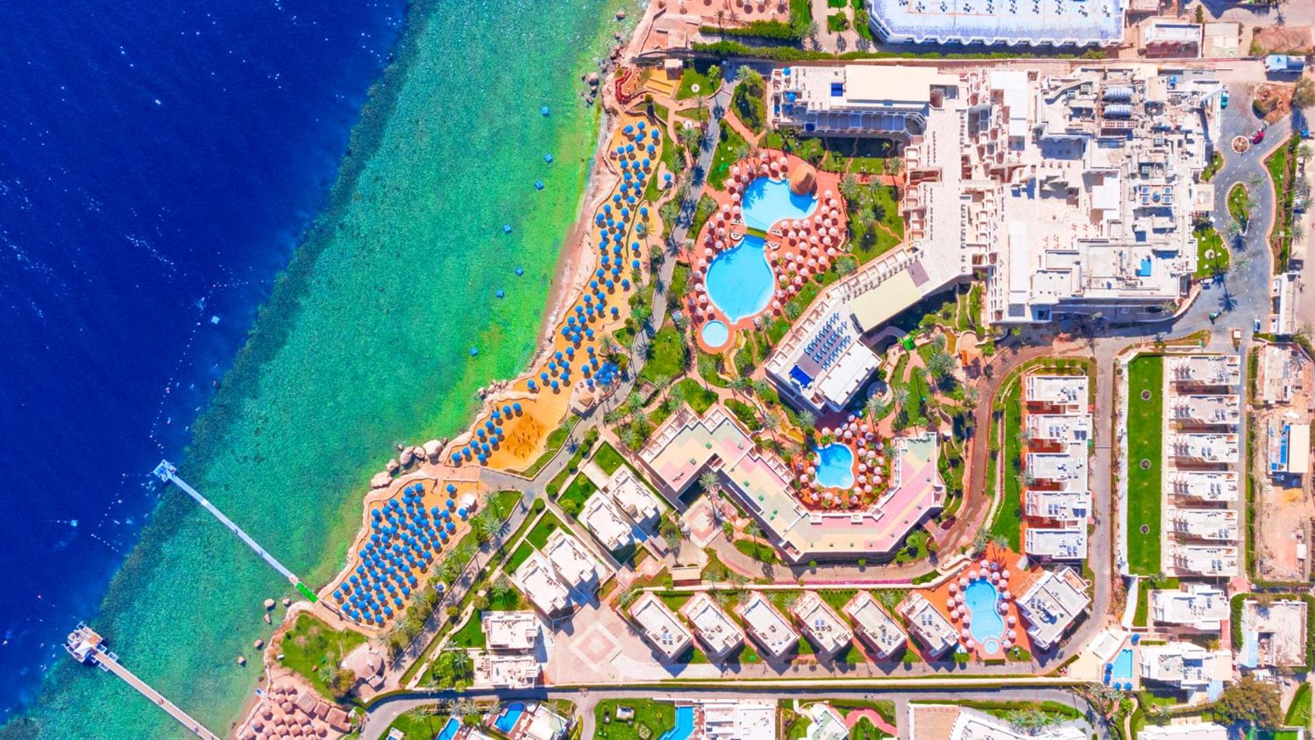 Pickalbatros Royal Grand Sharm - Adults Friendly 16 Years Plus Exterior photo The image shows an aerial view of a coastal resort area. It features various swimming pools outlined by landscaped gardens and pathways. There are also rows of sun loungers and umbrellas near the beach. The water transitions from a deep blue to a vib
