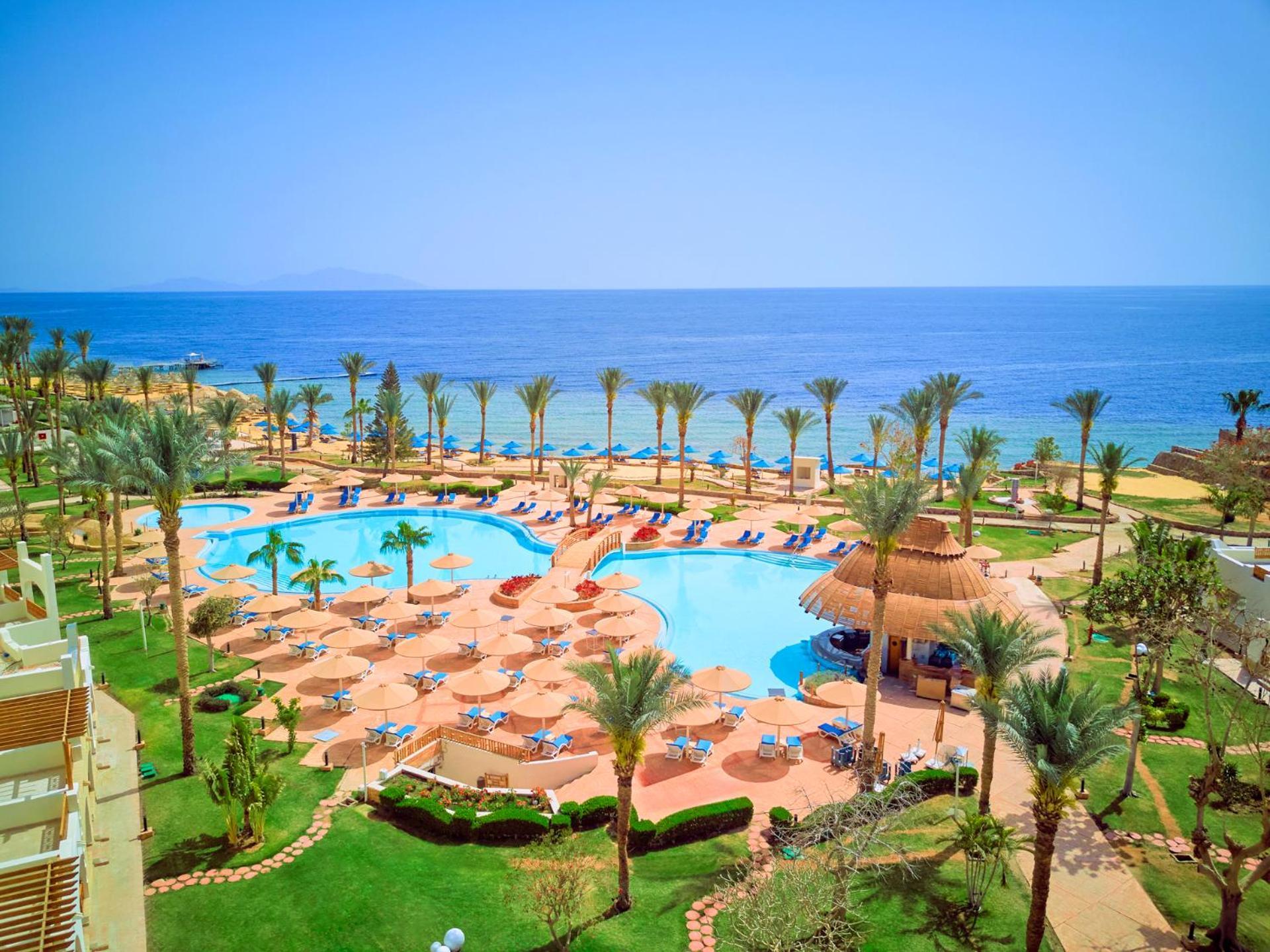 Pickalbatros Royal Grand Sharm - Adults Friendly 16 Years Plus Exterior photo The photo depicts a beautiful resort scene by the ocean. In the foreground, there is a large swimming pool surrounded by palm trees and lounge chairs. The pool area features sunbeds and umbrellas, suggesting a relaxing atmosphere for guests. In the b