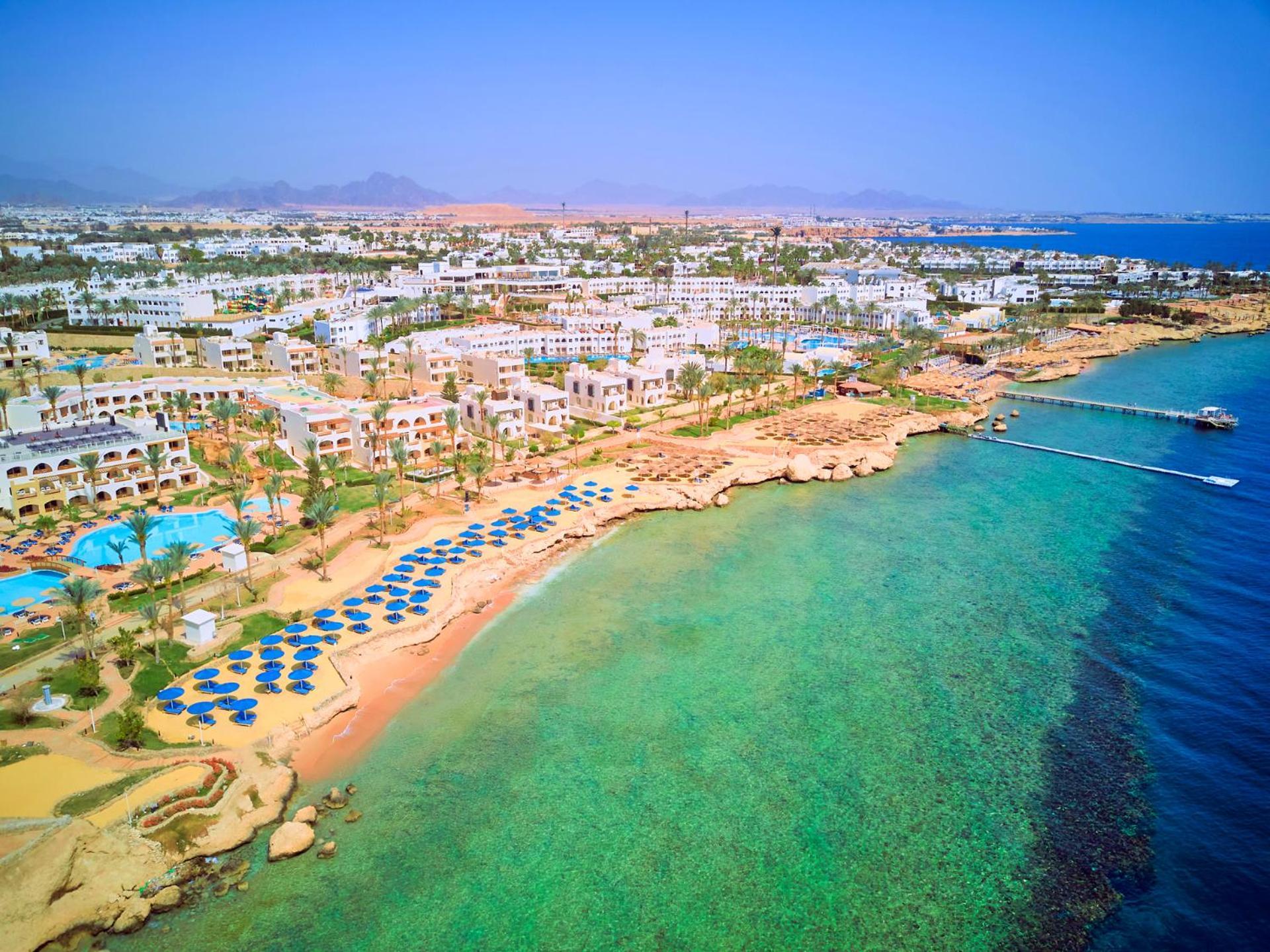 Pickalbatros Royal Grand Sharm - Adults Friendly 16 Years Plus Exterior photo The photo showcases a picturesque coastal scene, likely from a resort area. It features a sandy beach lined with blue umbrellas and lounge chairs, indicating a recreational space for relaxation. The water is clear and exhibits shades of green and blu