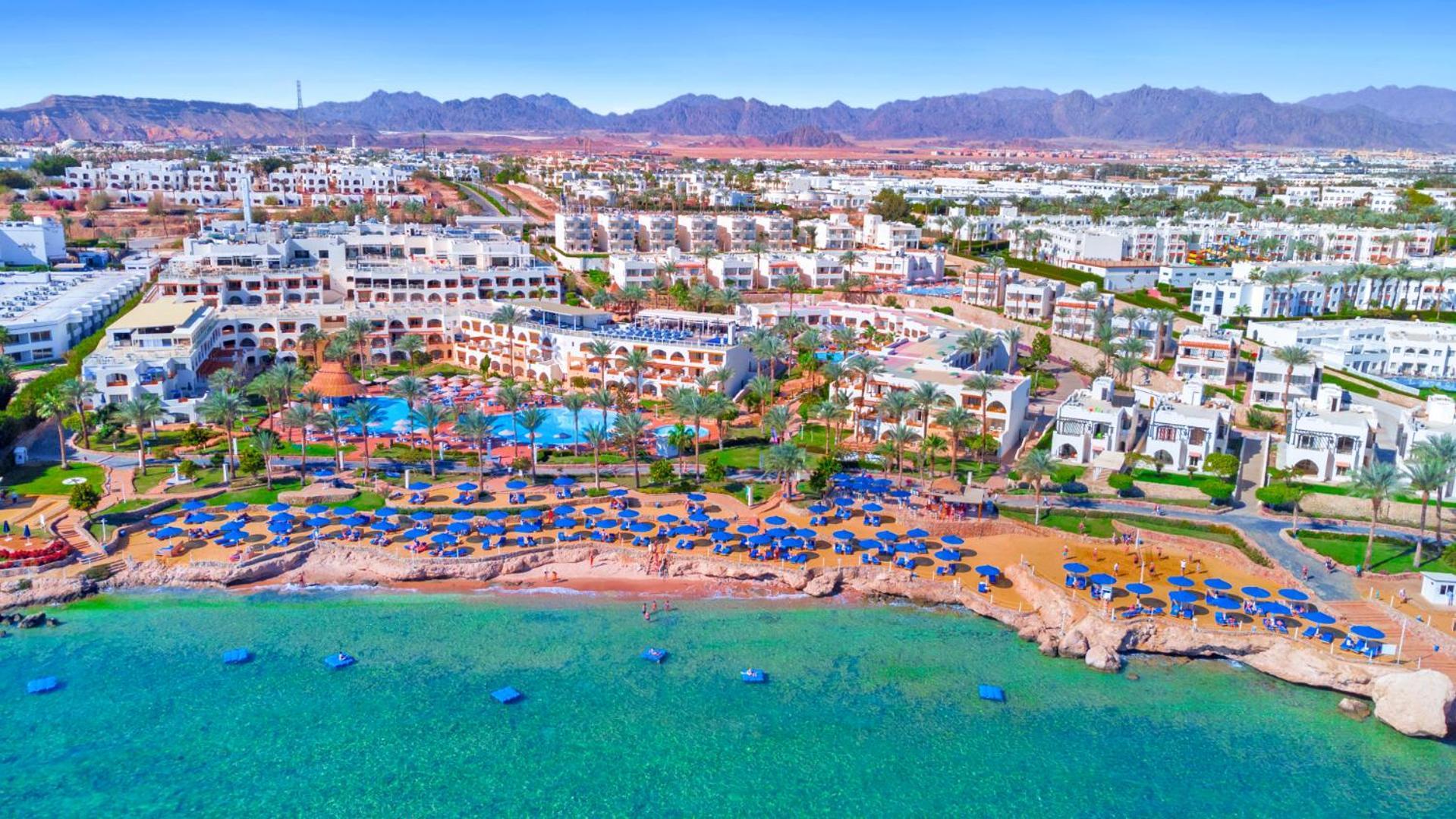 Pickalbatros Royal Grand Sharm - Adults Friendly 16 Years Plus Exterior photo The photo showcases a beautiful coastal resort area. In the foreground, there is a sandy beach lined with blue umbrellas and sun loungers. The vibrant turquoise water gently laps at the shore. Behind the beach, you can see a well-maintained resort wi