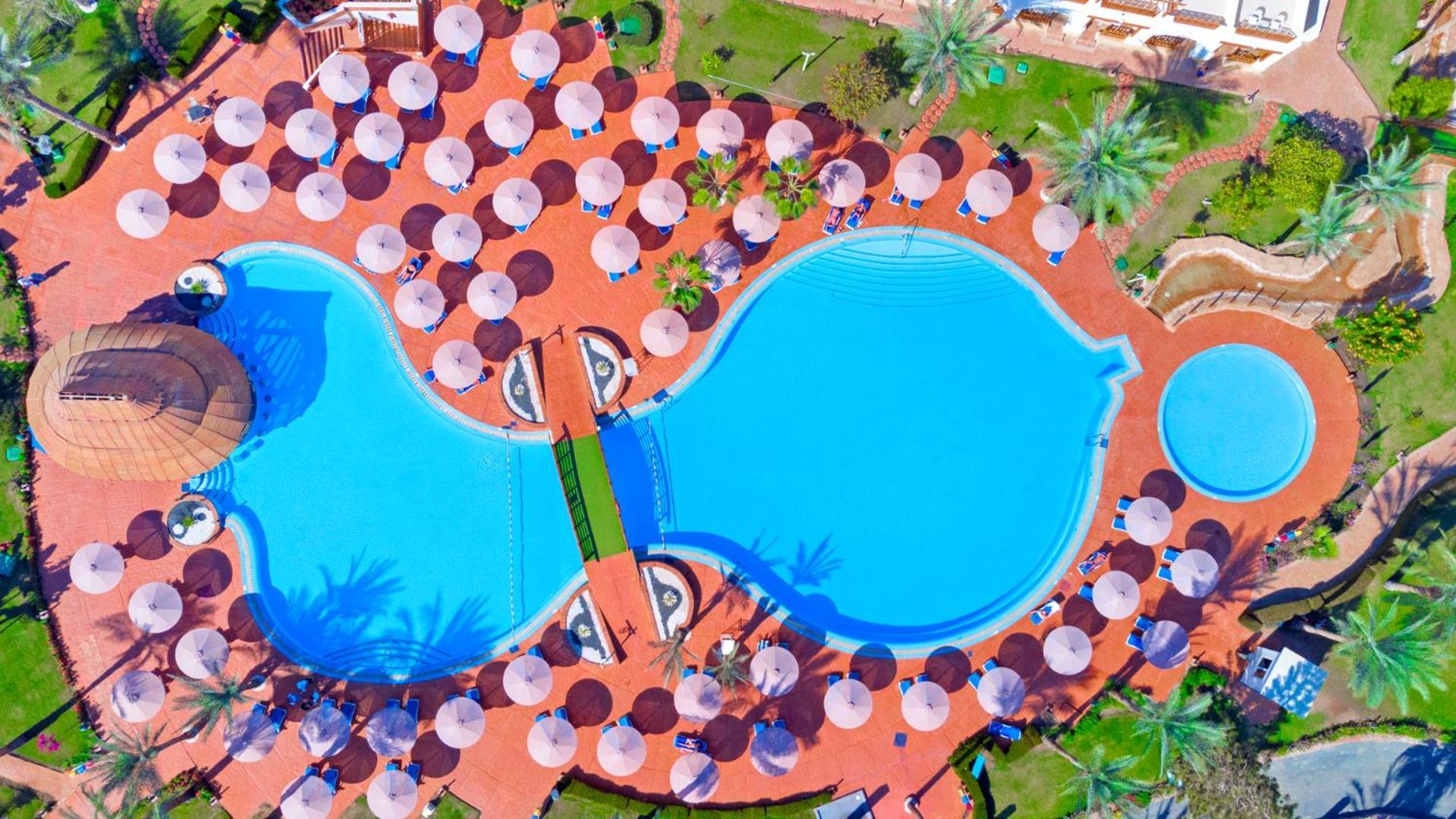 Pickalbatros Royal Grand Sharm - Adults Friendly 16 Years Plus Exterior photo The photo shows an aerial view of a large swimming pool area, shaped in a distinctive layout resembling two interconnected pools. Surrounding the main pool are numerous umbrellas, providing shade for visitors. The pool area is set on a reddish-colore