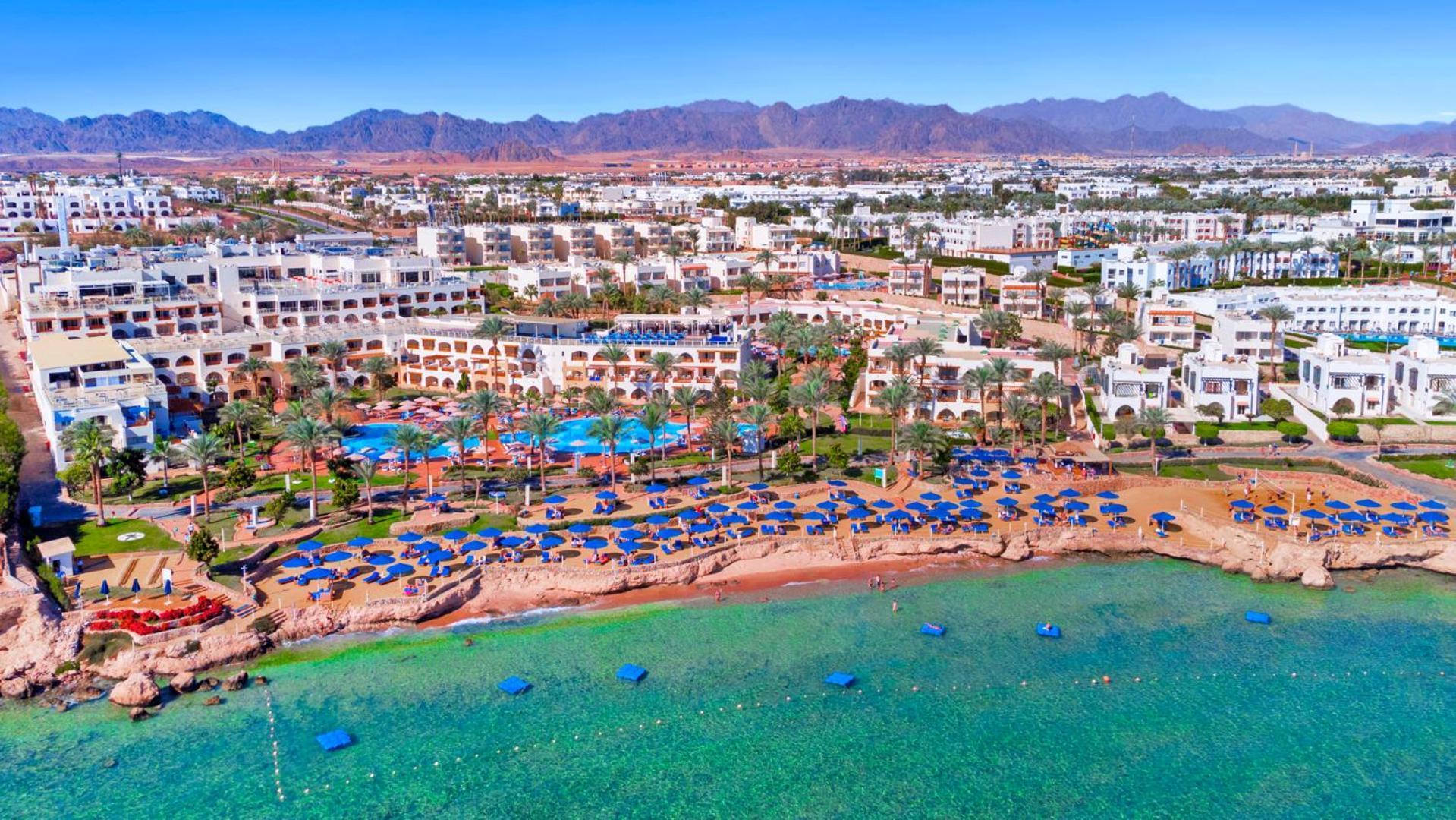 Pickalbatros Royal Grand Sharm - Adults Friendly 16 Years Plus Exterior photo The photo shows an aerial view of a coastal resort area. In the foreground, there is a beach with greenish-blue water, where several blue beach umbrellas and lounges are set up for guests. The shoreline appears sandy, and there are some rocky areas. 