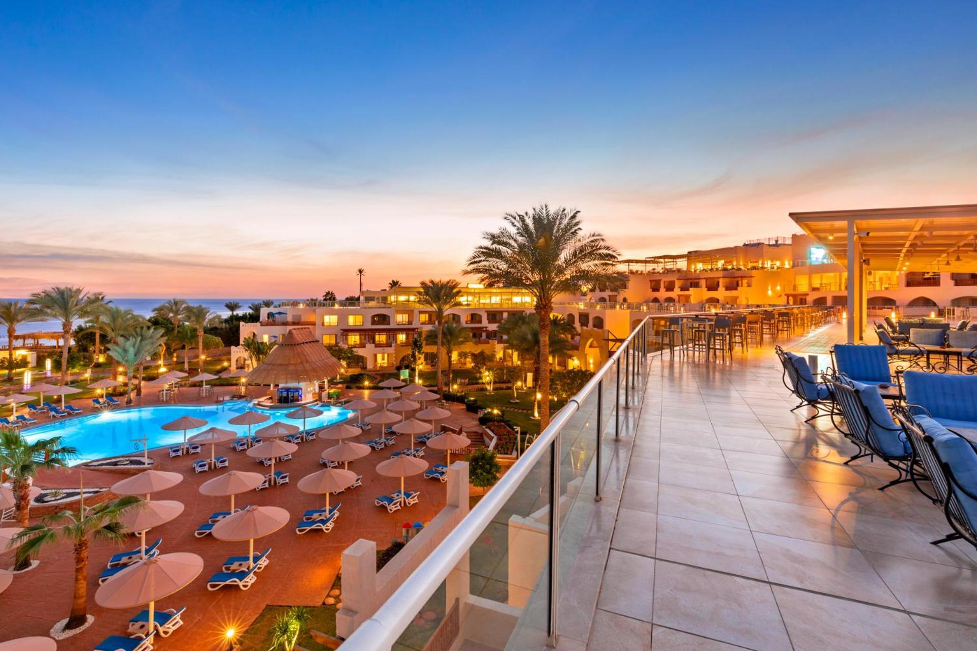 Pickalbatros Royal Grand Sharm - Adults Friendly 16 Years Plus Exterior photo The photo depicts a scenic view of a resort during sunset. In the foreground, there is a balcony with tiled flooring, overlooking the resort. Below, a swimming pool is visible, surrounded by lounge chairs and large umbrellas, indicating a relaxing at