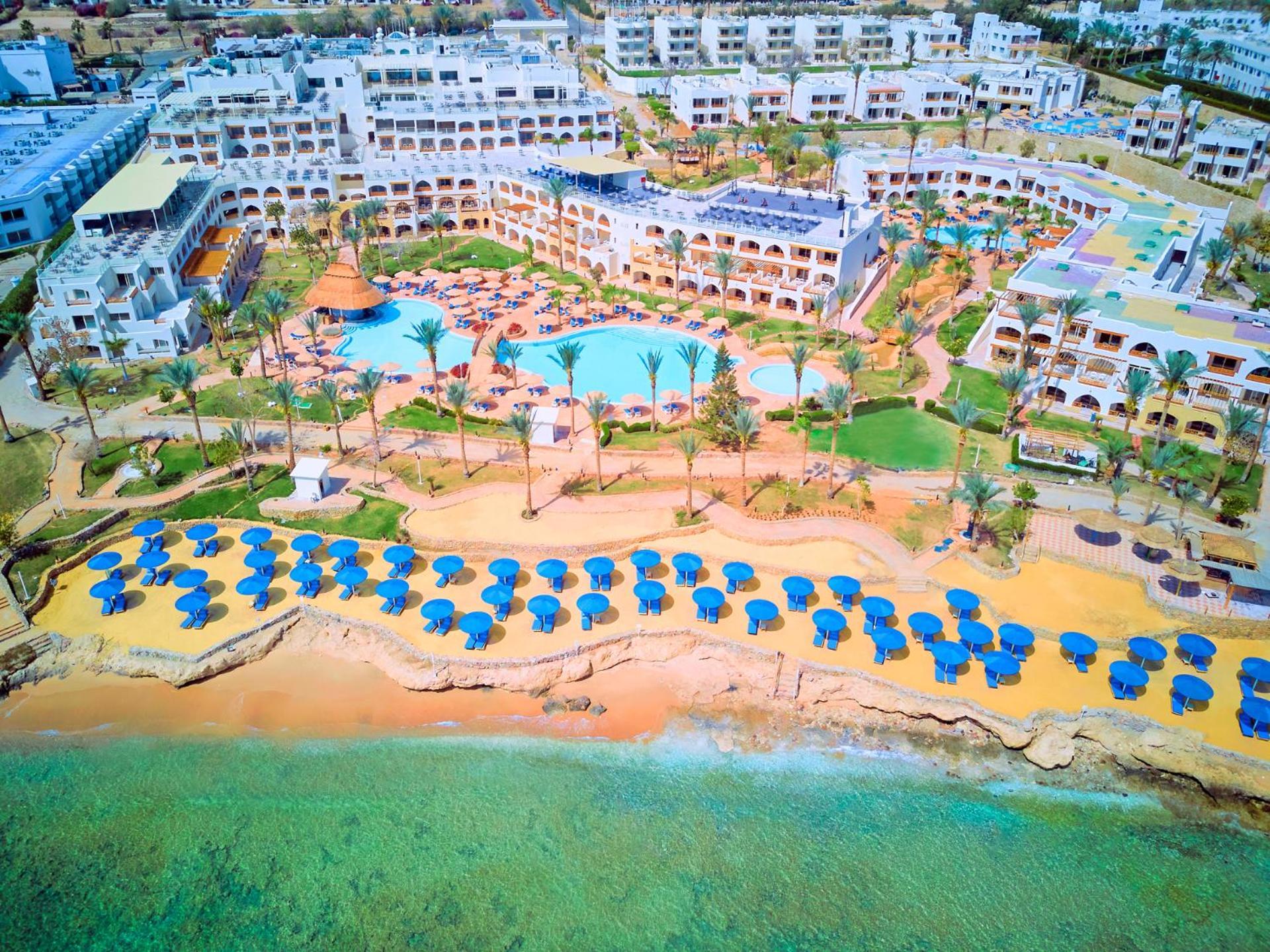 Pickalbatros Royal Grand Sharm - Adults Friendly 16 Years Plus Exterior photo The photo shows a large resort complex situated near a beach. There is a turquoise swimming pool at the center, surrounded by green lawns and palm trees. Sun loungers with blue umbrellas are lined up on the sandy beach, and the beach area is adjacent