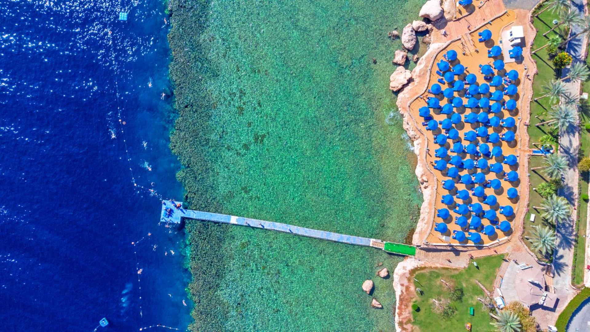 Pickalbatros Royal Grand Sharm - Adults Friendly 16 Years Plus Exterior photo The photo shows an aerial view of a beach area. In the foreground, there is a wooden pier extending into the clear blue water. To the right, there are numerous beach umbrellas with blue canopies arranged neatly, likely indicating a resort or beach cl