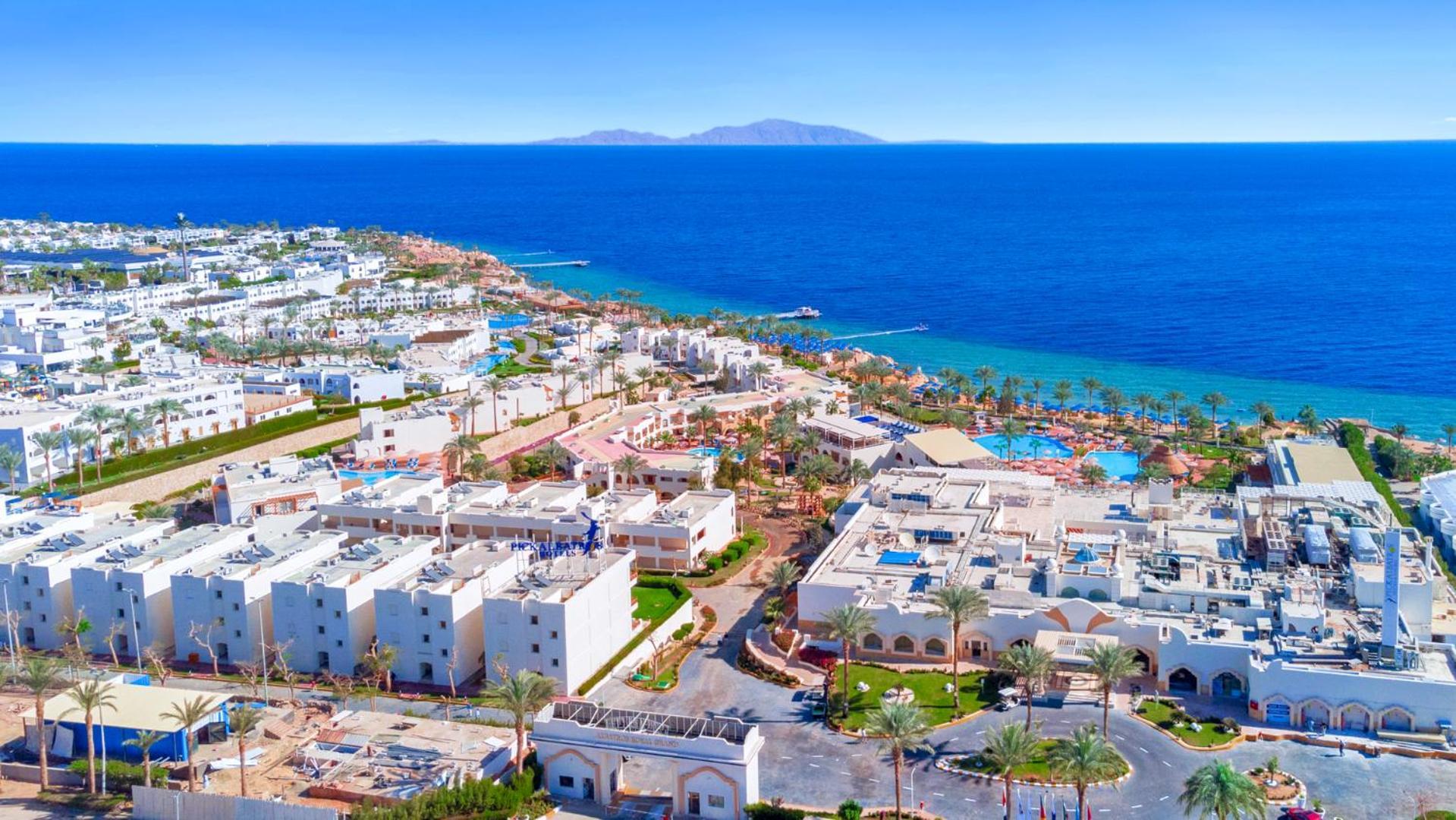 Pickalbatros Royal Grand Sharm - Adults Friendly 16 Years Plus Exterior photo The photo shows a beautiful coastal landscape, featuring a resort area by the sea. There are white buildings, likely hotels or villas, arranged in a sprawling layout near the beach. The water is a vibrant blue, contrasting with the sandy shore and gr