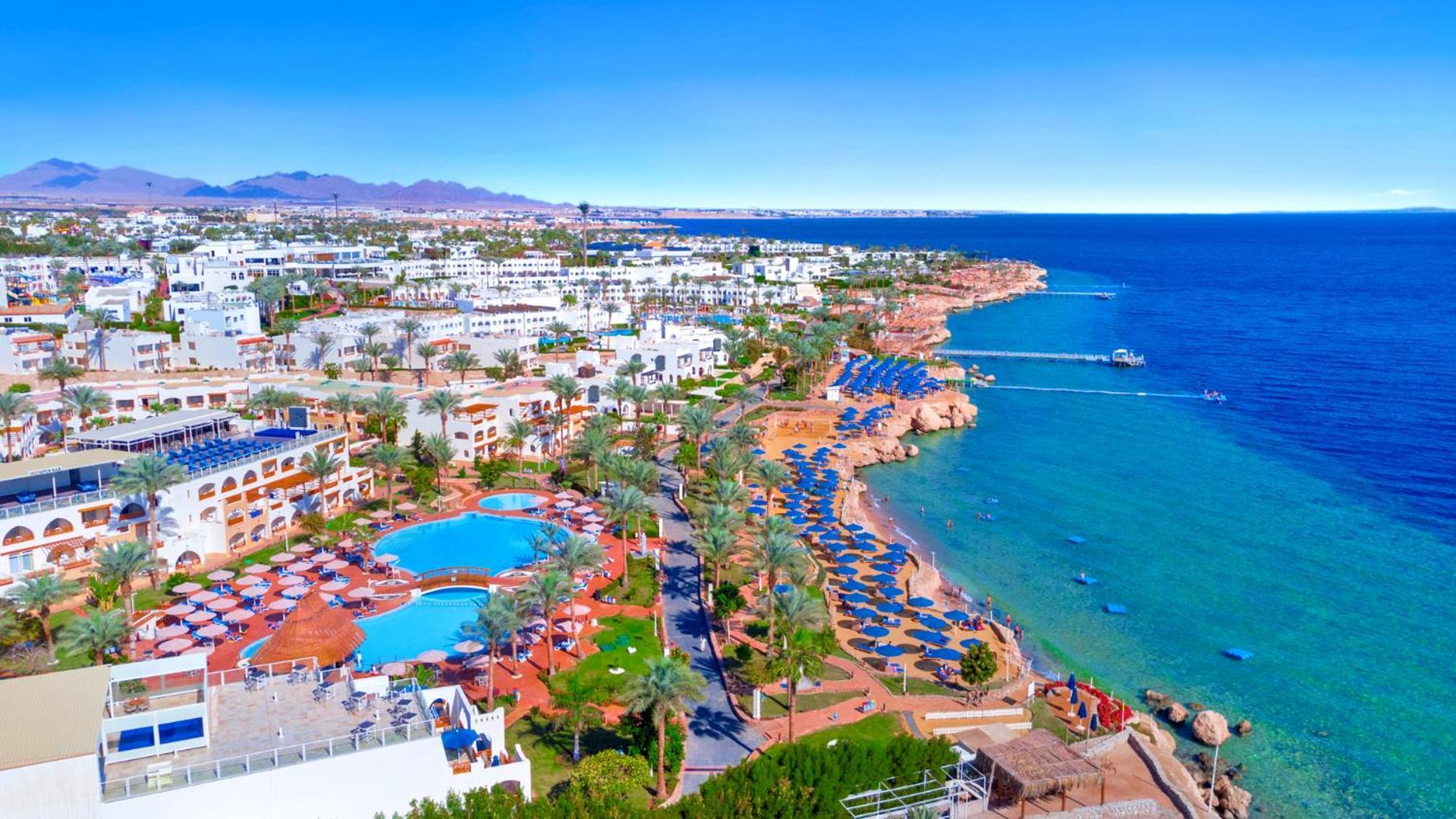 Pickalbatros Royal Grand Sharm - Adults Friendly 16 Years Plus Exterior photo The photo features a picturesque coastal scene, likely from a resort area. In the foreground, there is a large swimming pool surrounded by lounge chairs. Further back, the landscape includes several buildings, possibly hotels or apartments, that over