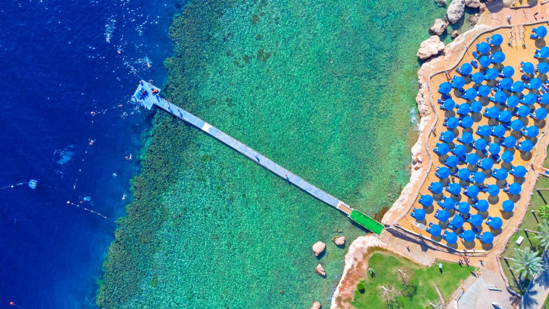 Pickalbatros Royal Grand Sharm - Adults Friendly 16 Years Plus Exterior photo The photo shows an aerial view of a coastal area featuring a long wooden pier extending into clear turquoise water. On one side of the pier, there appears to be a beach with umbrellas or sunbeds arranged neatly. The water is vibrant and visually invi