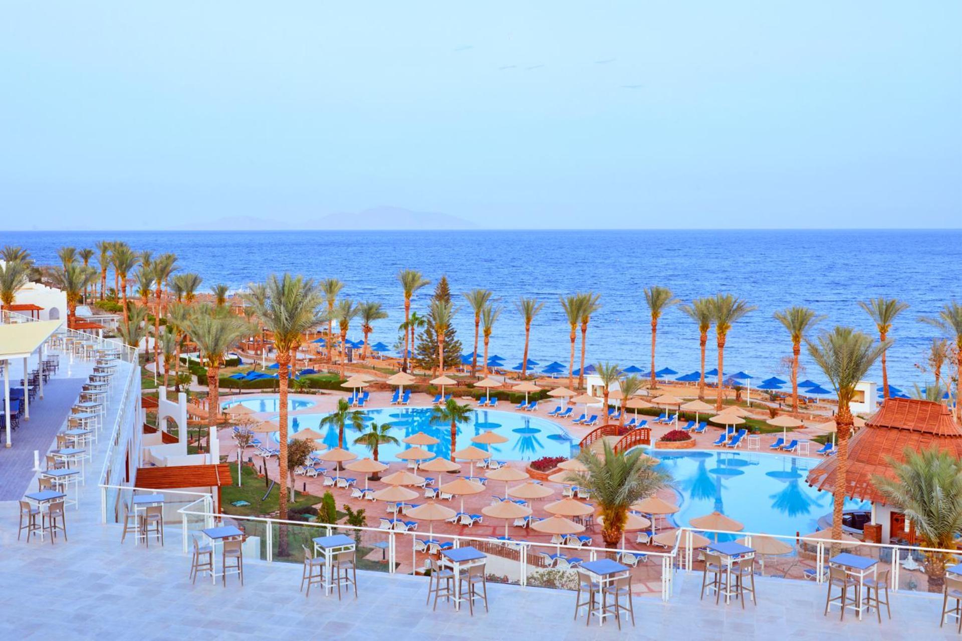 Pickalbatros Royal Grand Sharm - Adults Friendly 16 Years Plus Exterior photo The photo depicts a scenic resort area by the sea. In the foreground, there is a large swimming pool surrounded by lounge chairs and umbrellas. Palm trees line the sides of the pool, adding to the tropical atmosphere. In the background, a beautiful b