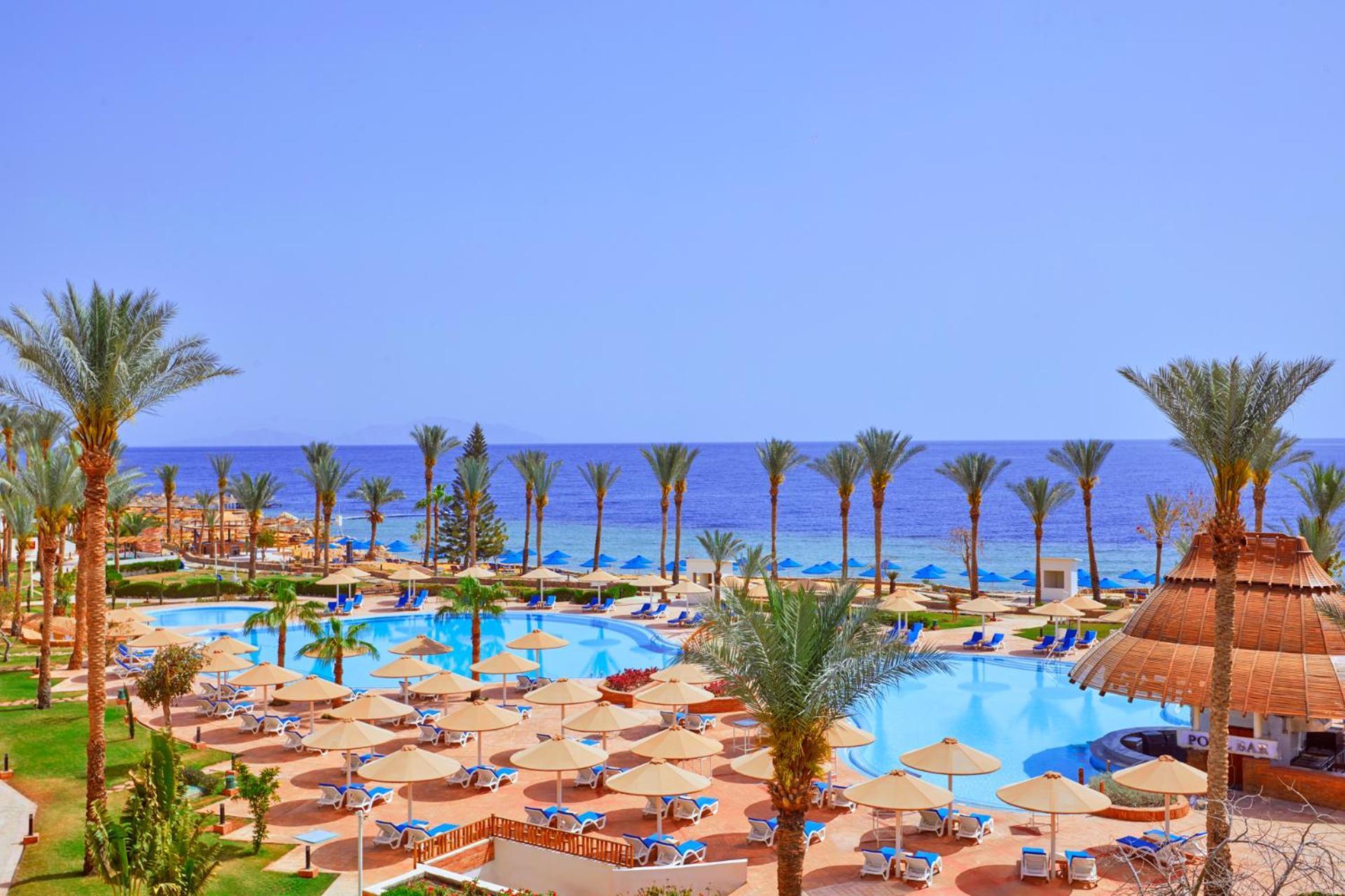 Pickalbatros Royal Grand Sharm - Adults Friendly 16 Years Plus Exterior photo The photo showcases a beautiful resort setting by the sea. It features a large swimming pool surrounded by sunny lounge chairs and umbrellas, perfect for relaxation. The area is adorned with lush palm trees, adding to the tropical vibe. In the backgr