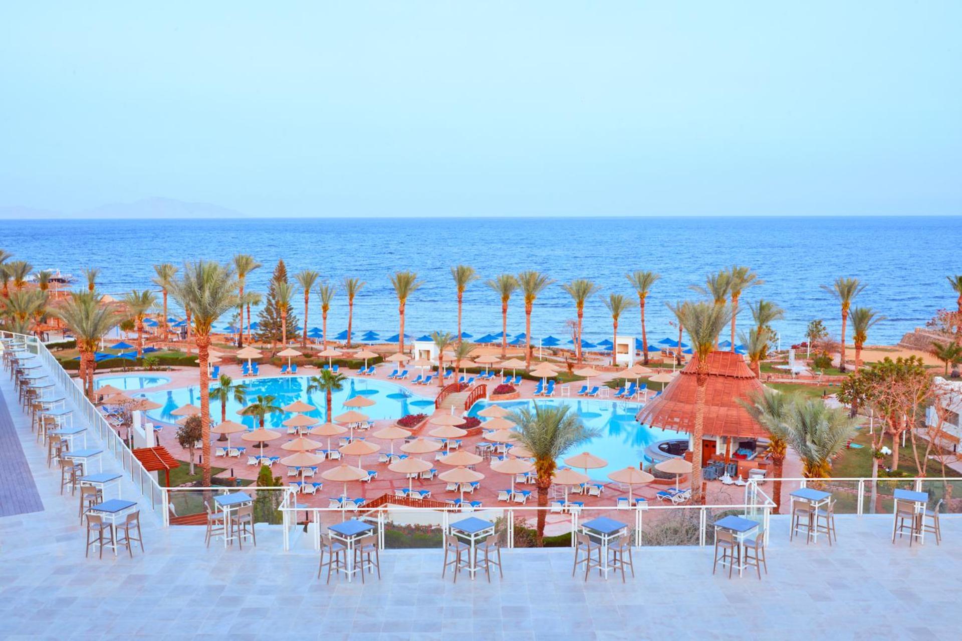Pickalbatros Royal Grand Sharm - Adults Friendly 16 Years Plus Exterior photo The photo shows a scenic view of a resort or beach area. In the foreground, there are several seating areas with bar stools on a terrace. Below, a large swimming pool is visible, surrounded by lounging chairs and umbrellas. Palm trees line the area, 