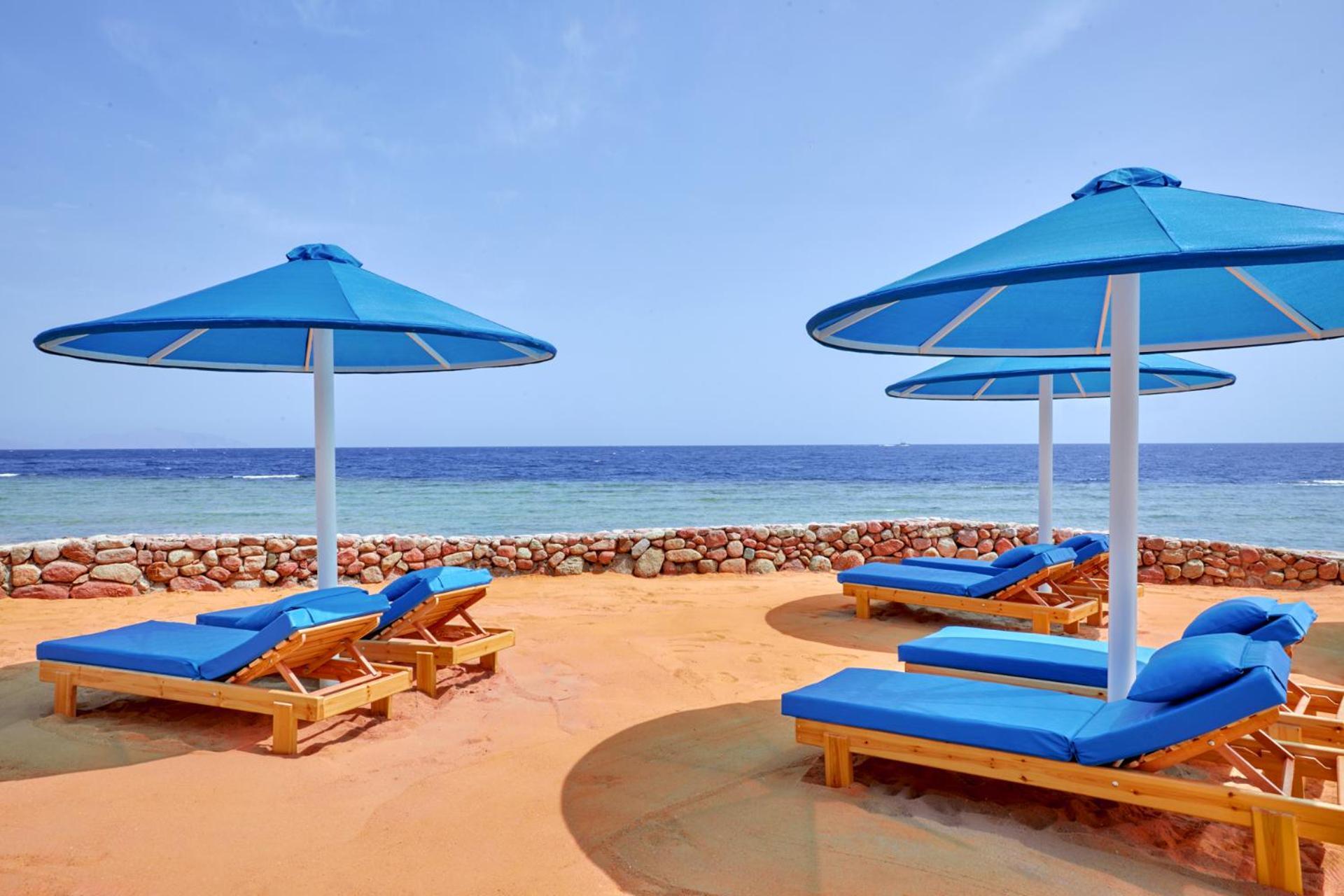 Pickalbatros Royal Grand Sharm - Adults Friendly 16 Years Plus Exterior photo The photo shows a beach scene featuring several blue umbrellas and lounge chairs on a sandy area. The umbrellas are large and have a bright blue color, providing shade for the lounge chairs underneath. In the background, there's a view of the ocean w