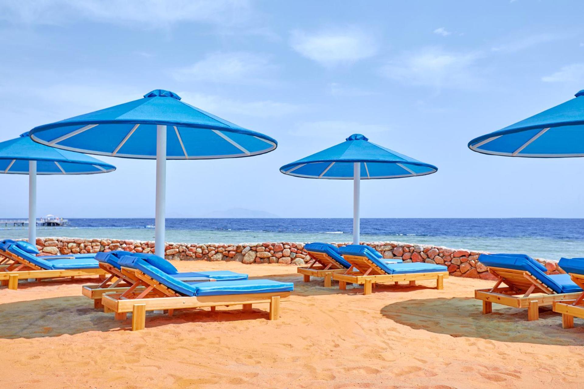 Pickalbatros Royal Grand Sharm - Adults Friendly 16 Years Plus Exterior photo The photo shows a beach scene featuring several blue umbrellas providing shade over wooden lounge chairs. The sandy beach extends toward a calm sea, creating a relaxing atmosphere. The sky is mostly clear, with a few clouds, suggesting pleasant weath