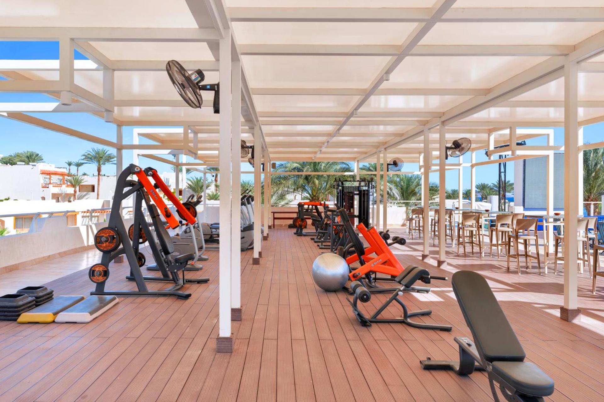 Pickalbatros Royal Grand Sharm - Adults Friendly 16 Years Plus Exterior photo The photo depicts a gym area located outdoors or in a covered terrace, featuring various fitness equipment. There are several weight machines and benches, as well as some free weights. Large fans are installed for ventilation, and the space is design