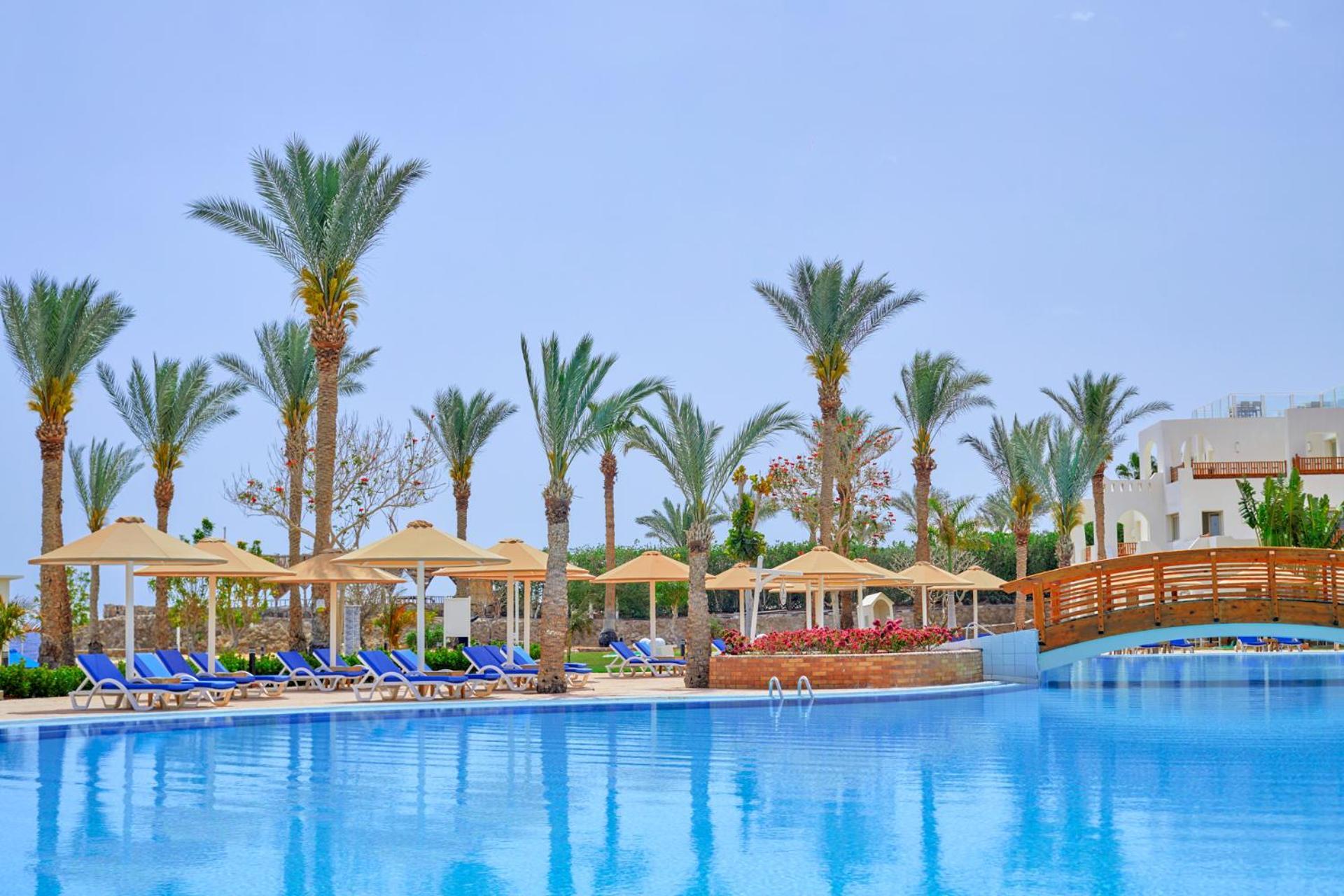 Pickalbatros Royal Grand Sharm - Adults Friendly 16 Years Plus Exterior photo The photo shows a serene poolside scene at a resort. There is a large, clear blue pool reflecting the sky, surrounded by lounge chairs with blue cushions. Palm trees line the area, adding a tropical feel. Several covered loungers are placed under umb