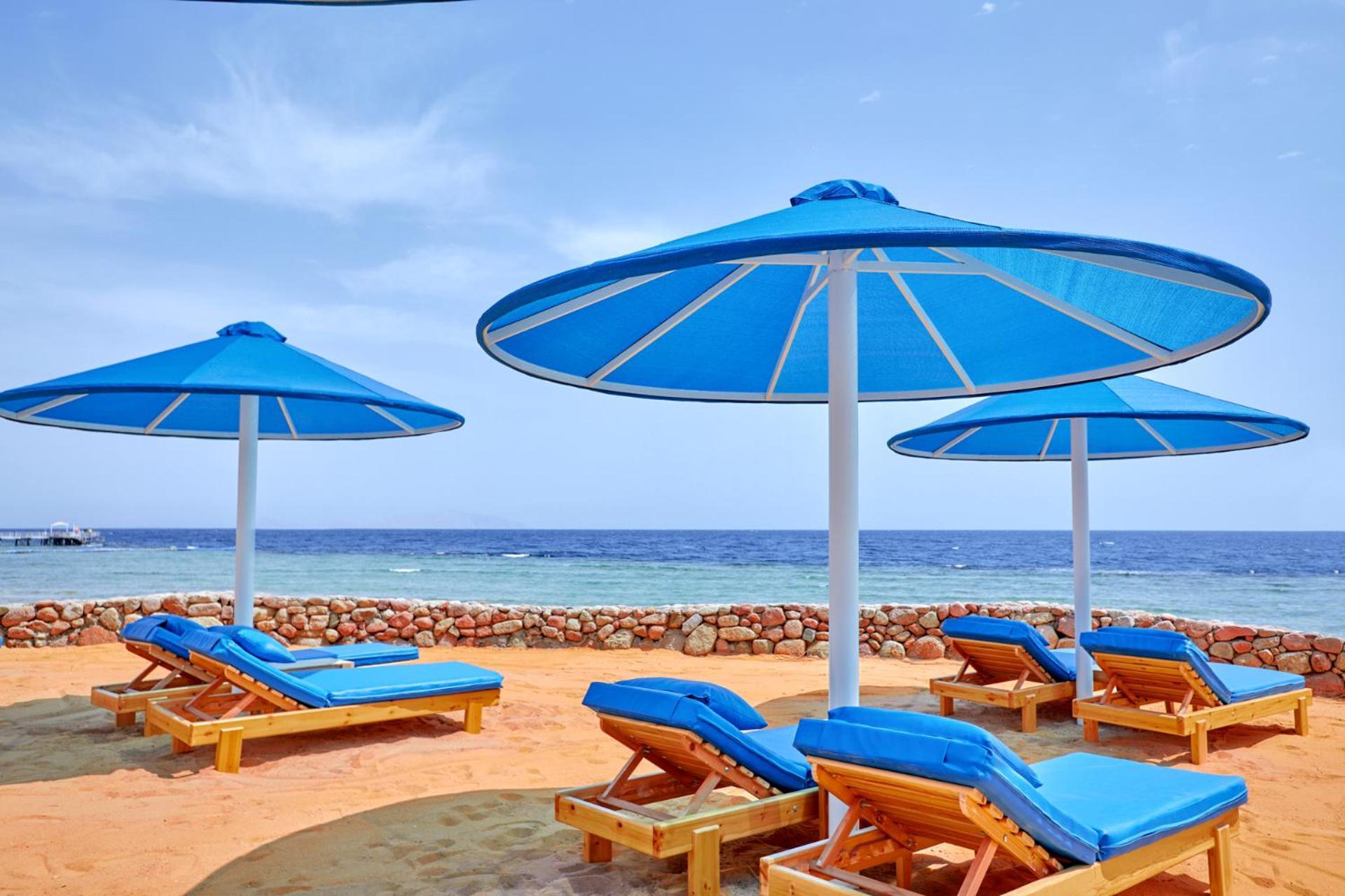 Pickalbatros Royal Grand Sharm - Adults Friendly 16 Years Plus Exterior photo The photo shows a serene beach scene with several blue sun loungers arranged beneath large blue umbrellas. The loungers are positioned on sandy ground, with the ocean visible in the background. The sky is clear, indicating a bright and sunny day, and