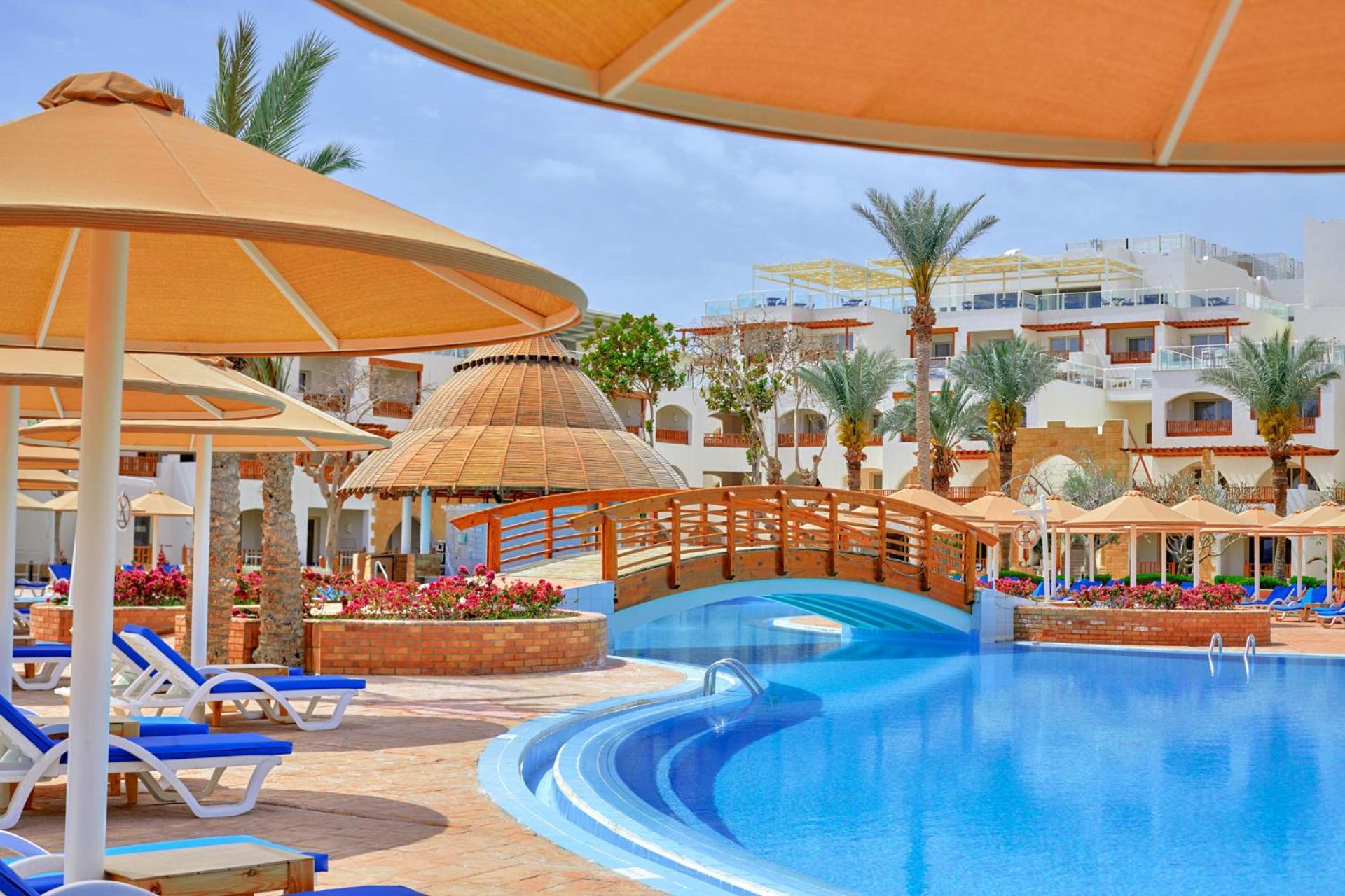 Pickalbatros Royal Grand Sharm - Adults Friendly 16 Years Plus Exterior photo The photo shows a picturesque resort scene featuring a bright blue swimming pool surrounded by sun loungers and parasols. In the background, there is a wooden bridge arching over a section of the pool, leading towards the other side. The area is ador