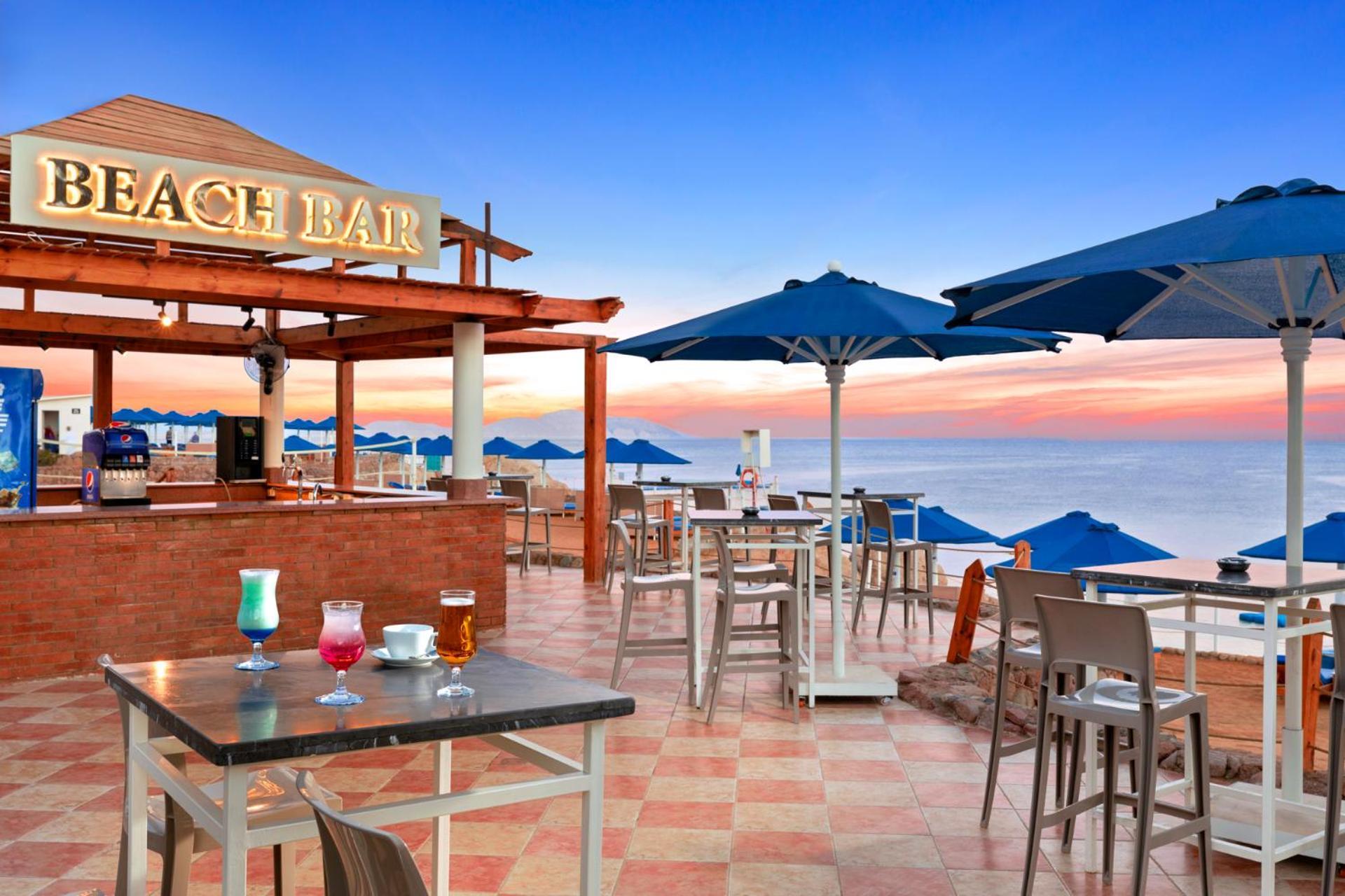 Pickalbatros Royal Grand Sharm - Adults Friendly 16 Years Plus Exterior photo The photo shows a beach bar setting, featuring a wooden structure with the words "BEACH BAR" prominently displayed. There are several tables and chairs arranged on a tiled floor, with some drinks placed on the tables. Blue umbrellas provide shade for