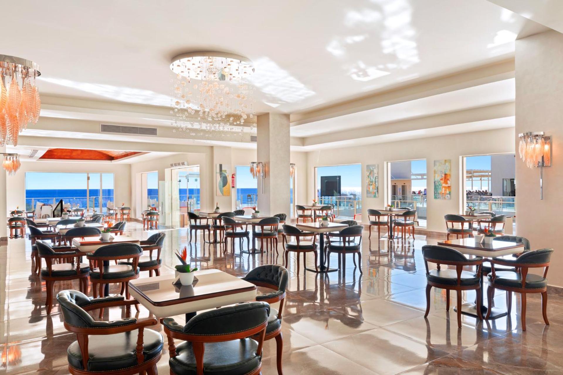 Pickalbatros Royal Grand Sharm - Adults Friendly 16 Years Plus Exterior photo The photo shows a bright and elegant restaurant interior. There are several tables arranged throughout the space, each with chairs around them. The decor features modern lighting, including a chandelier, and large windows that allow natural light to 