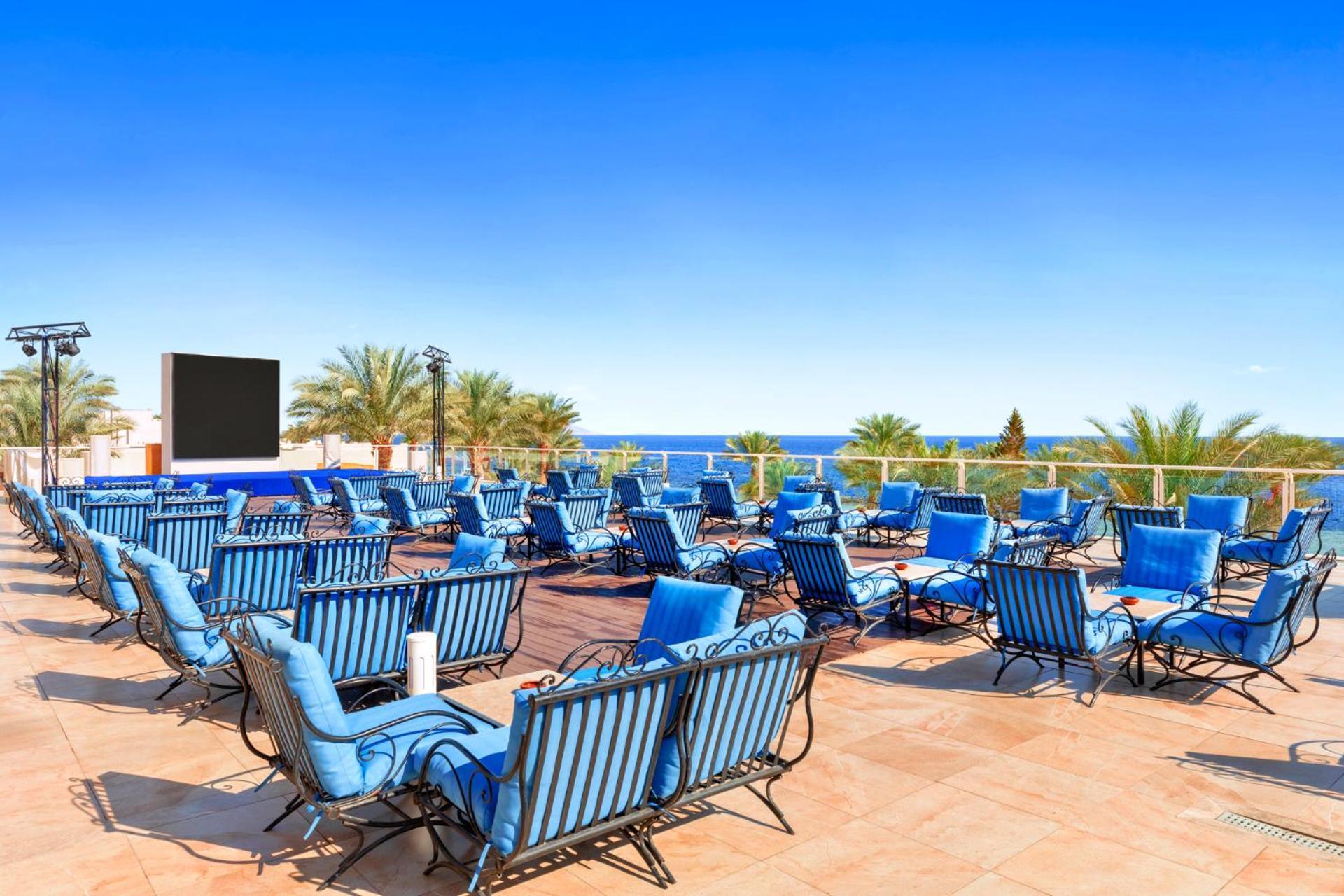 Pickalbatros Royal Grand Sharm - Adults Friendly 16 Years Plus Exterior photo The photo features a sunny outdoor space with numerous blue lounge chairs arranged in rows on a tiled patio. In the background, there is a clear blue sky and a view of the ocean, along with some palm trees adding to the tropical atmosphere. Additiona
