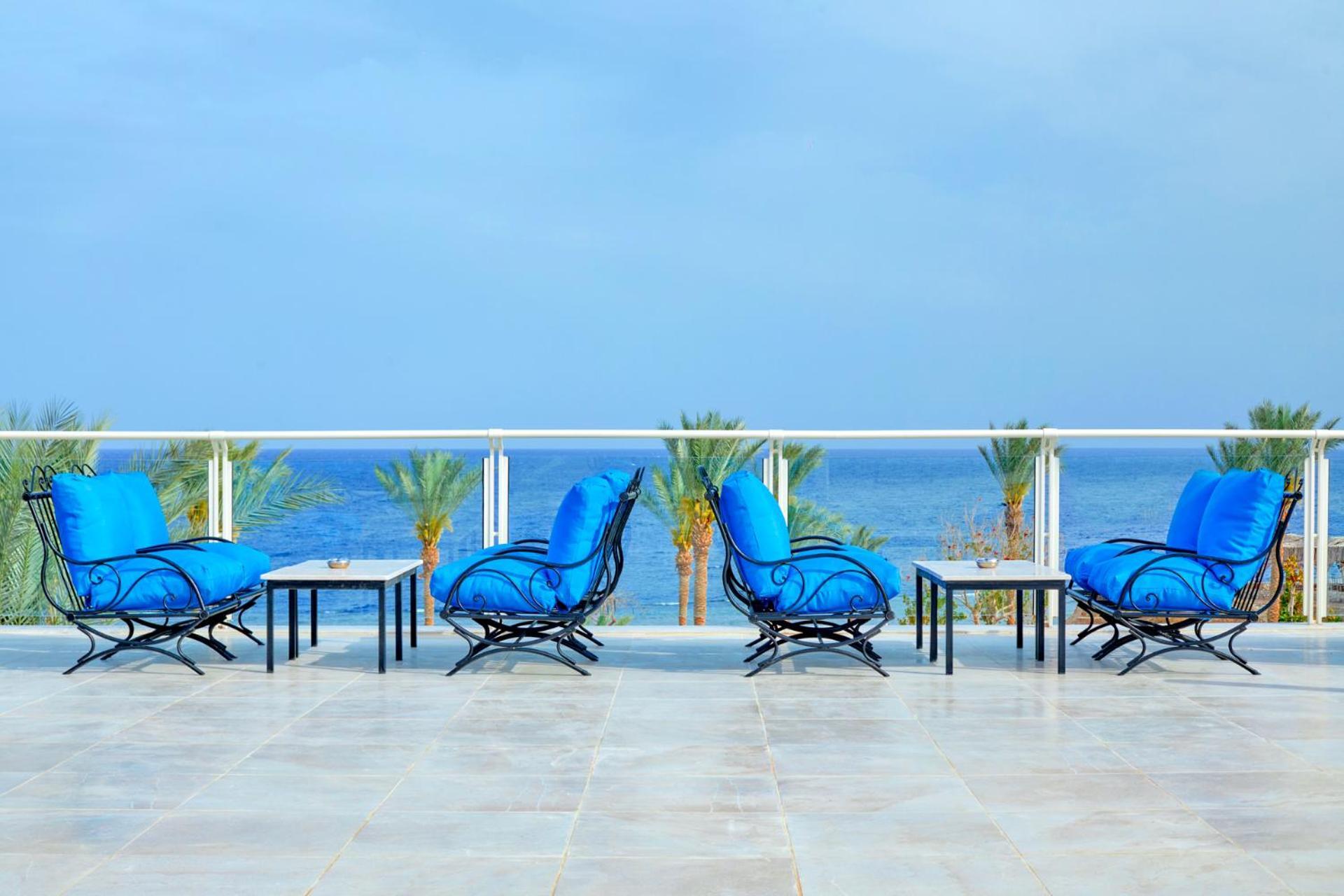 Pickalbatros Royal Grand Sharm - Adults Friendly 16 Years Plus Exterior photo The photo depicts a serene outdoor patio area with four blue lounge chairs arranged around small tables. The setting features a clear view of the ocean with palm trees visible in the background, complementing the tranquil atmosphere. The overall colo