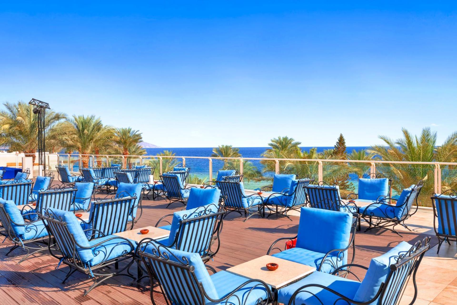 Pickalbatros Royal Grand Sharm - Adults Friendly 16 Years Plus Exterior photo The photo shows an outdoor deck or terrace area, adorned with numerous blue lounge chairs arranged neatly. In the background, there are palm trees and a clear blue sea that stretches toward the horizon under a bright sky. The setting appears to be a 