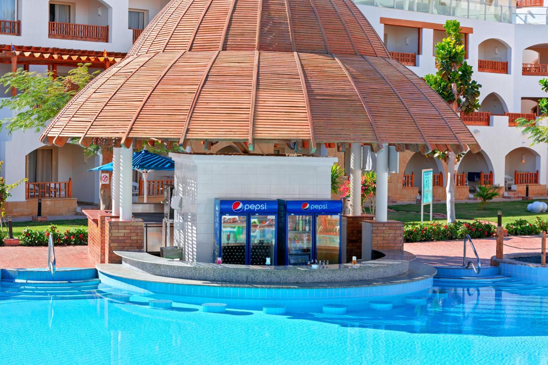 Pickalbatros Royal Grand Sharm - Adults Friendly 16 Years Plus Exterior photo The photo shows a poolside bar with a thatched roof structure, located near a swimming pool. The bar features a counter where drinks are served, and there are vending machines displaying the Pepsi brand. Surrounding the bar, there are neatly landscap